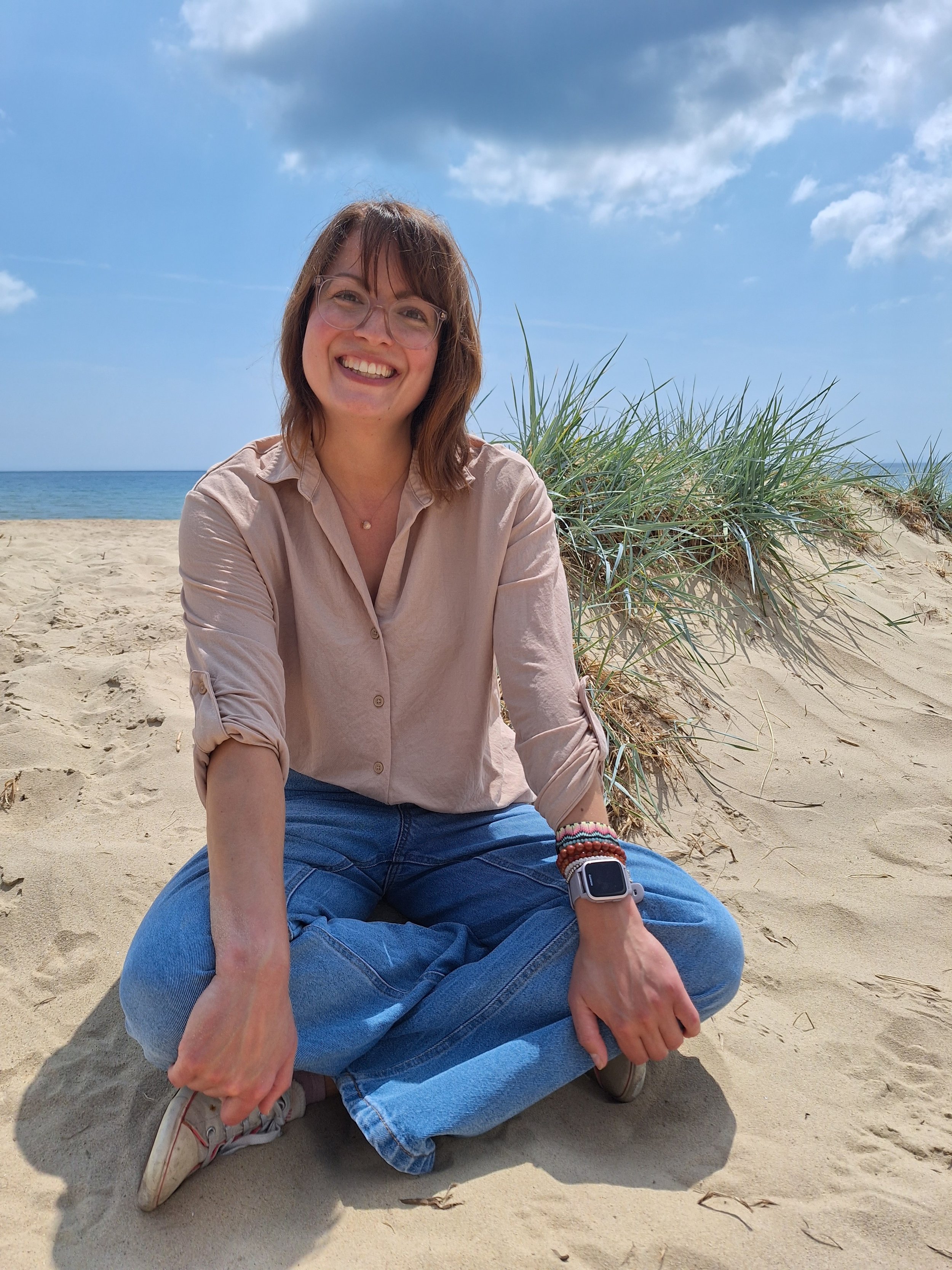 A woman with brown hair, glasses, and a friendly smile, sitting cross-legged on the sandy beach near some grass, with the ocean and a cloudy blue sky in the background.