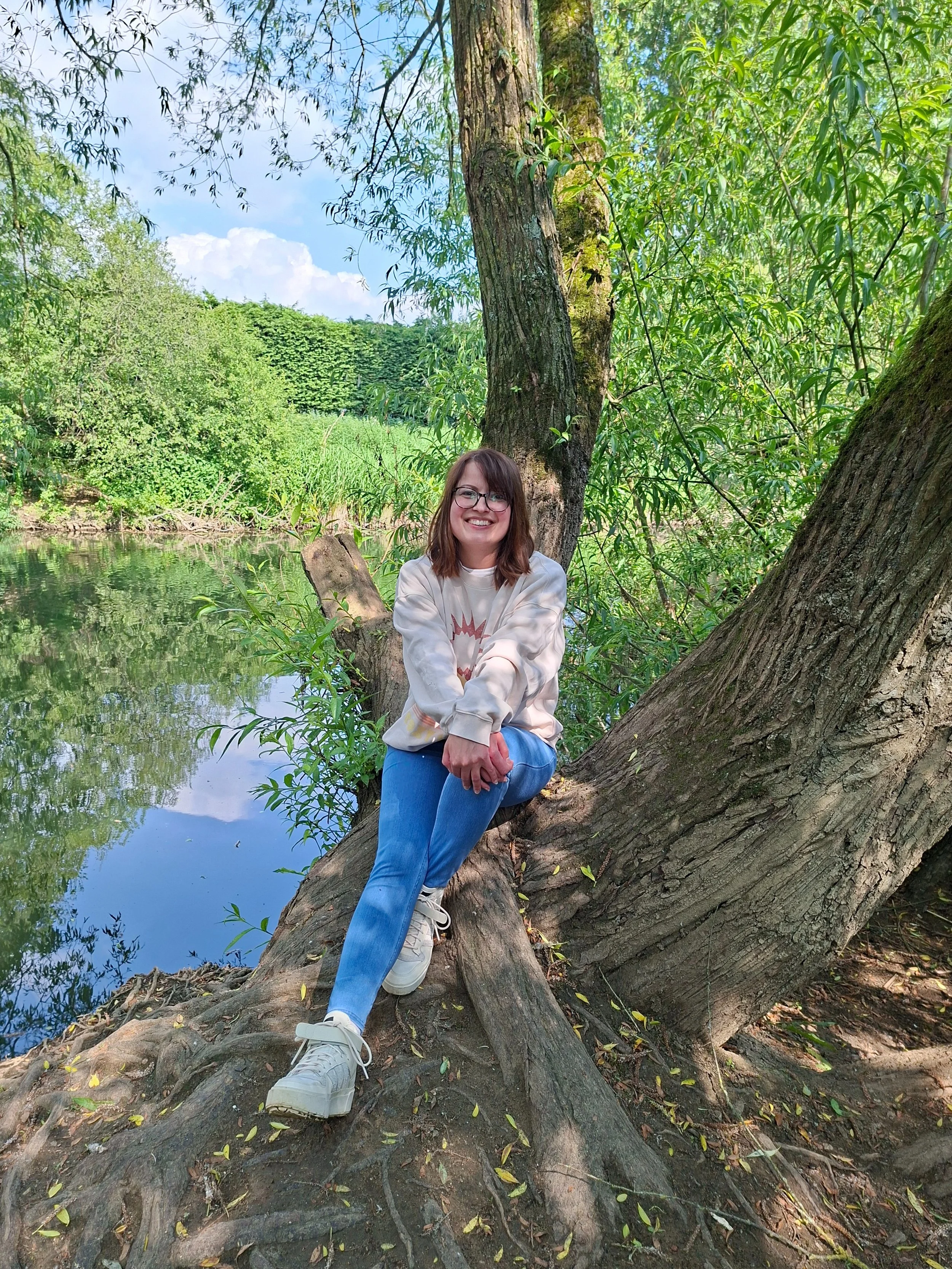 A young woman with glasses, wearing a light-colored sweatshirt and blue jeans, sitting on a tree root with a big tree behind her, in a lush green outdoor setting by a body of water.