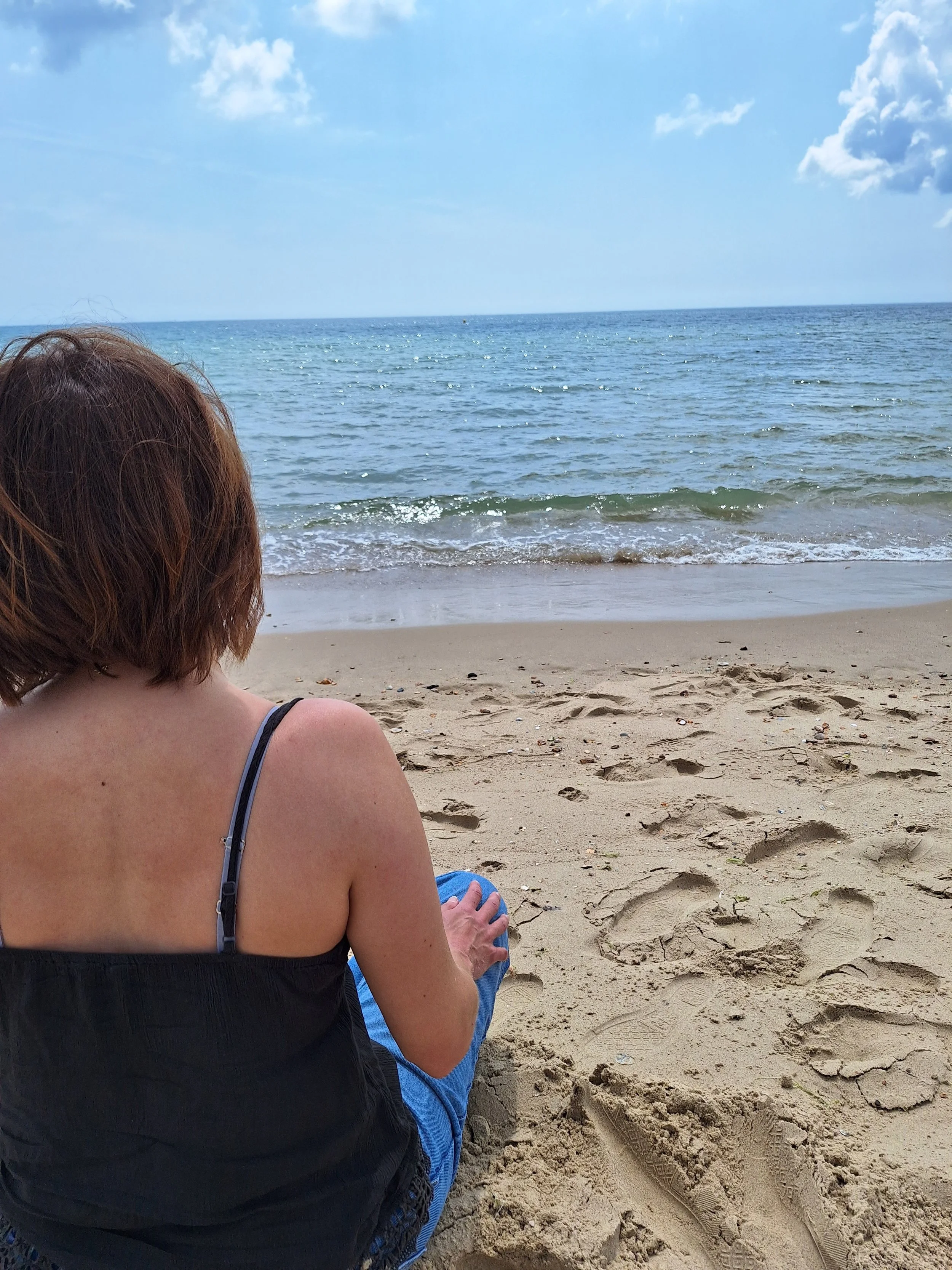 A woman with reddish-brown hair sitting on a sandy beach, facing the ocean under a partly cloudy sky.