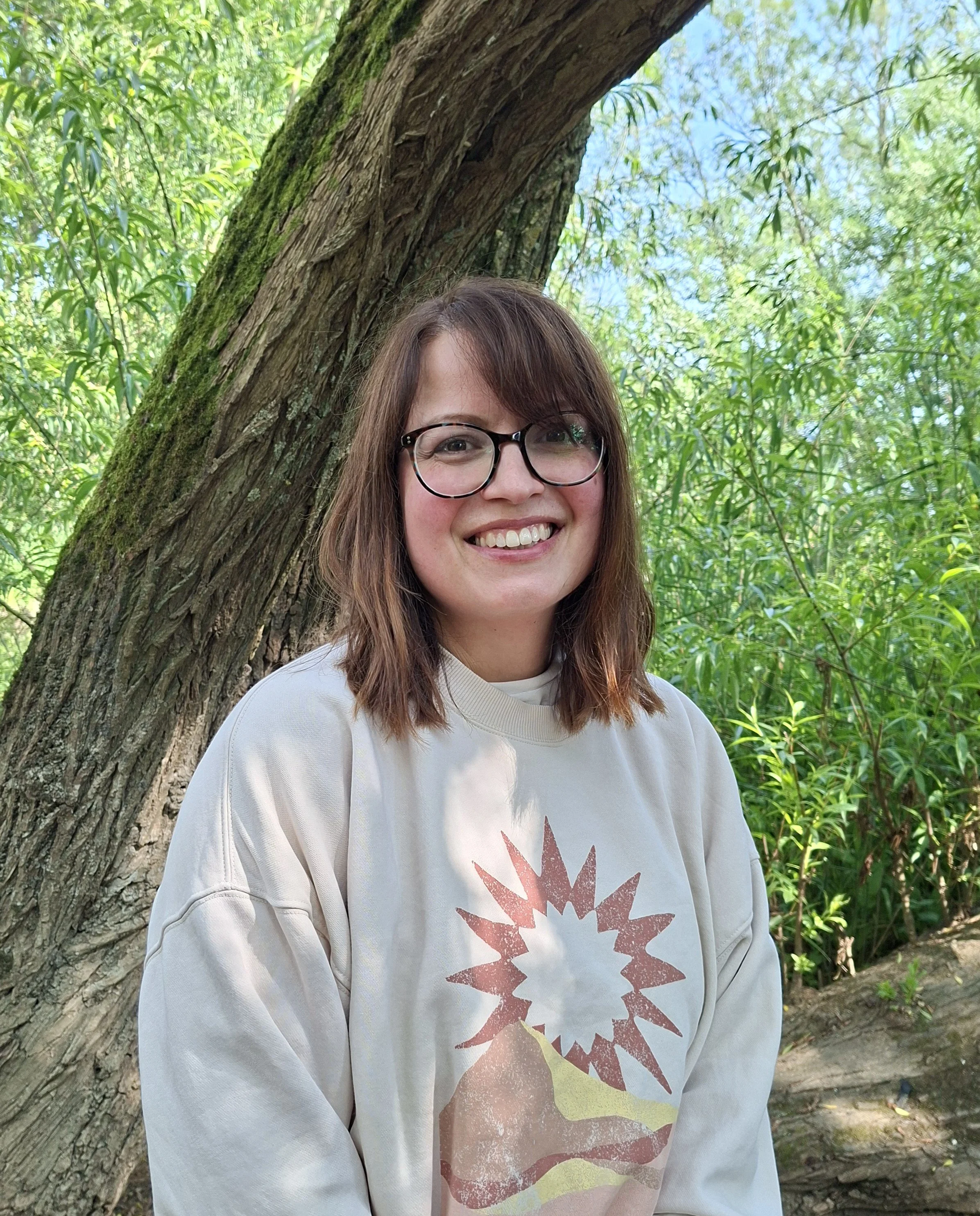 A smiling woman with glasses and brown hair standing outdoors near a large tree with green foliage.