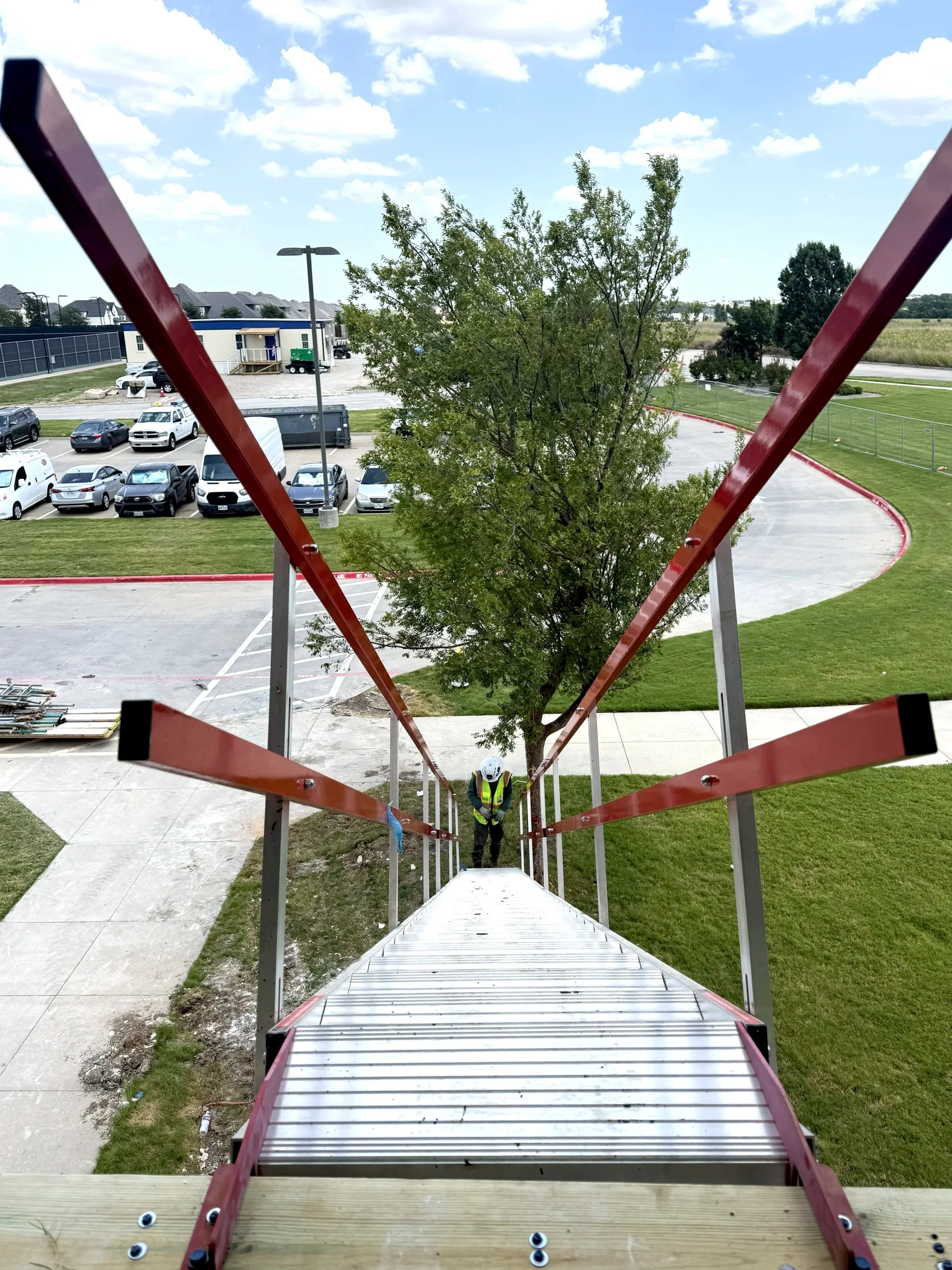 A worker in a safety vest and helmet standing at the bottom of a tall ladder, which is viewed from the top looking down. The ladder is red with black rubber feet, surrounded by grass, and a parking lot with several cars. A tree is in the background, 