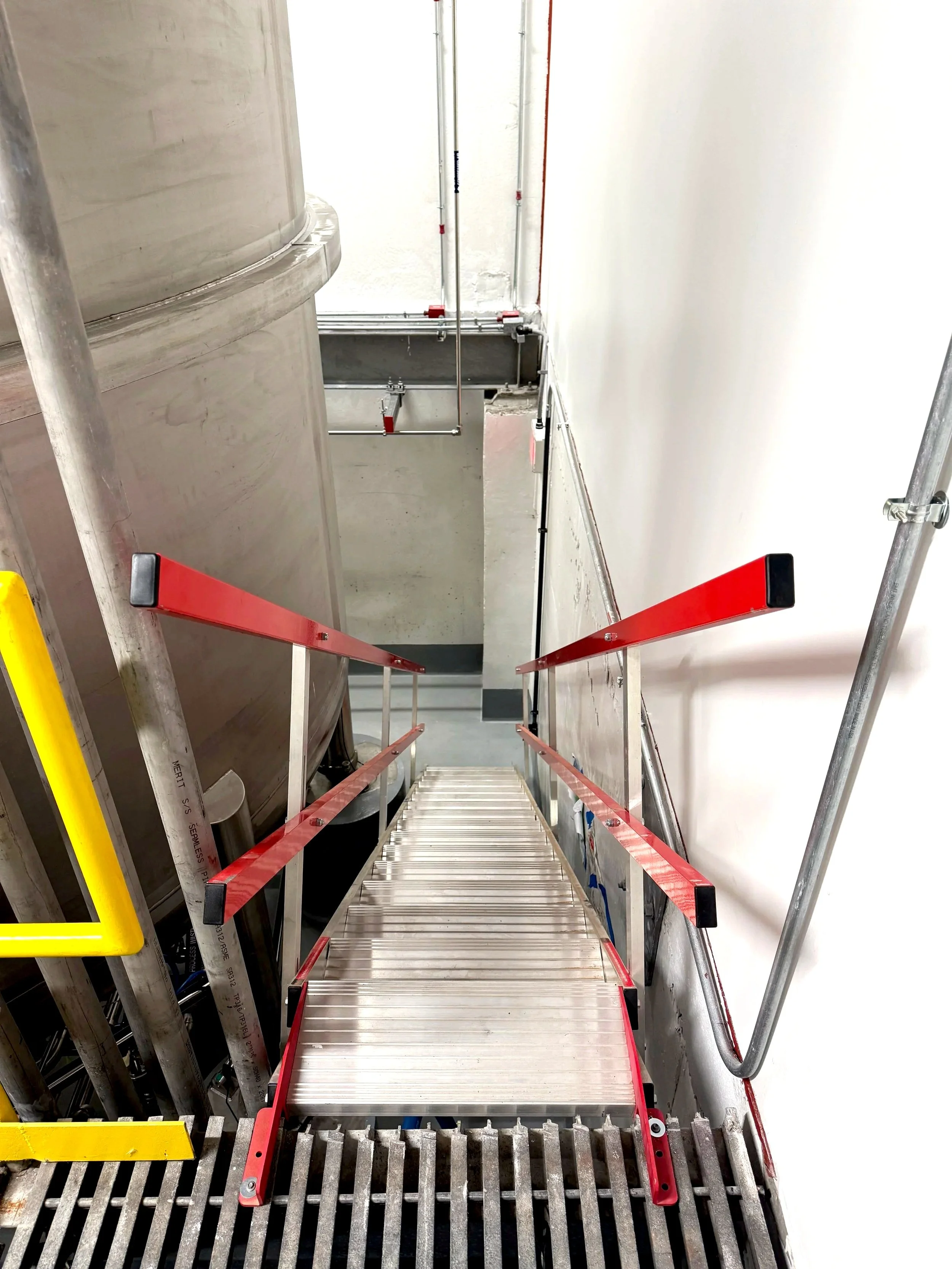 A construction staircase with red safety rails, metallic steps, and a metal grating at the bottom, leading down between unfinished concrete and drywall walls.