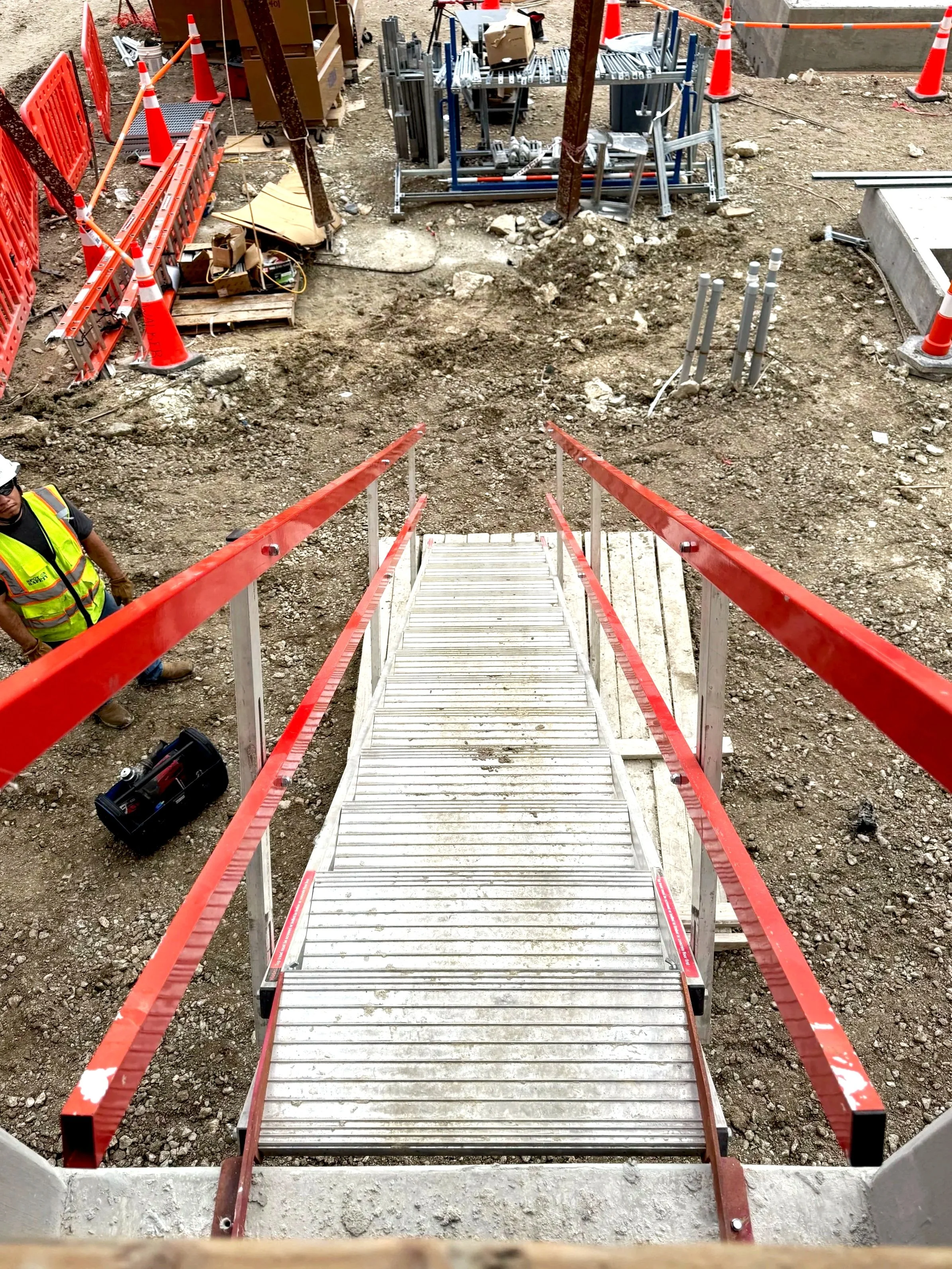 A construction site with a metal stairway leading down to an excavation area. Orange safety cones and barriers surround the site, with construction tools and materials scattered around. A worker wearing a yellow safety vest and helmet is partially vi