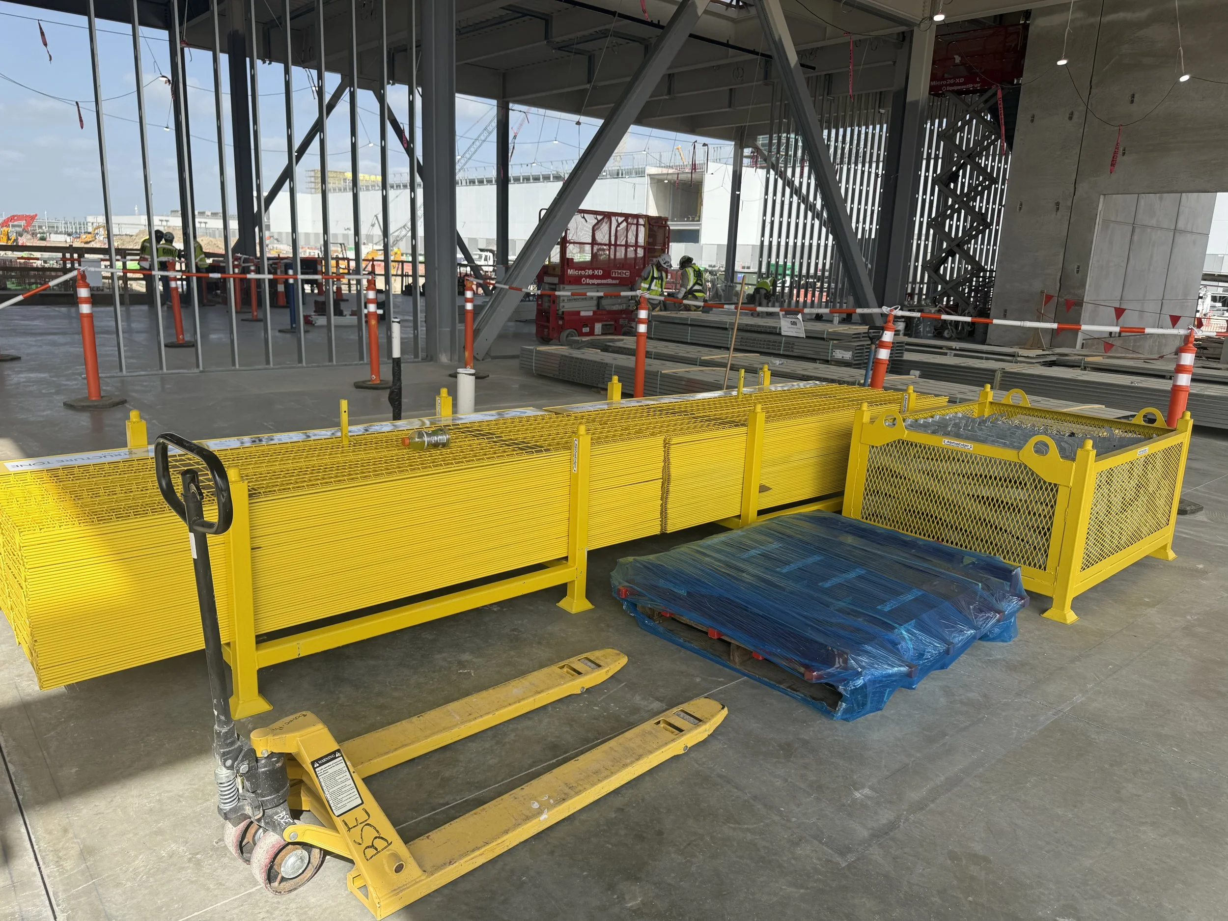 Construction site with workers in safety gear, orange and white cones, yellow safety rail, scaffolding, and building structures in progress.
