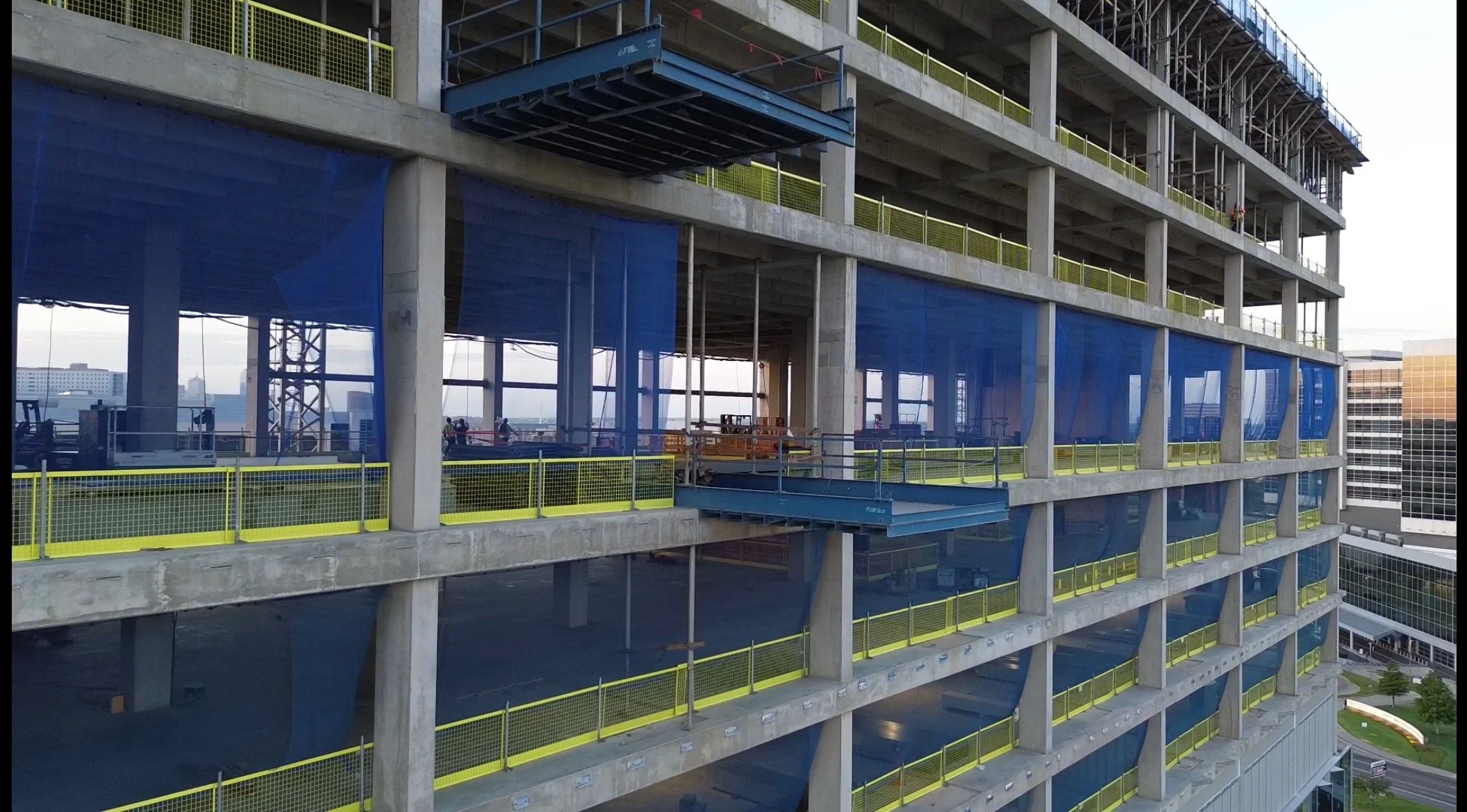 Side view of a high-rise building under construction with concrete floors, yellow safety railings, and blue safety nets on each level. Construction workers are visible on some floors.