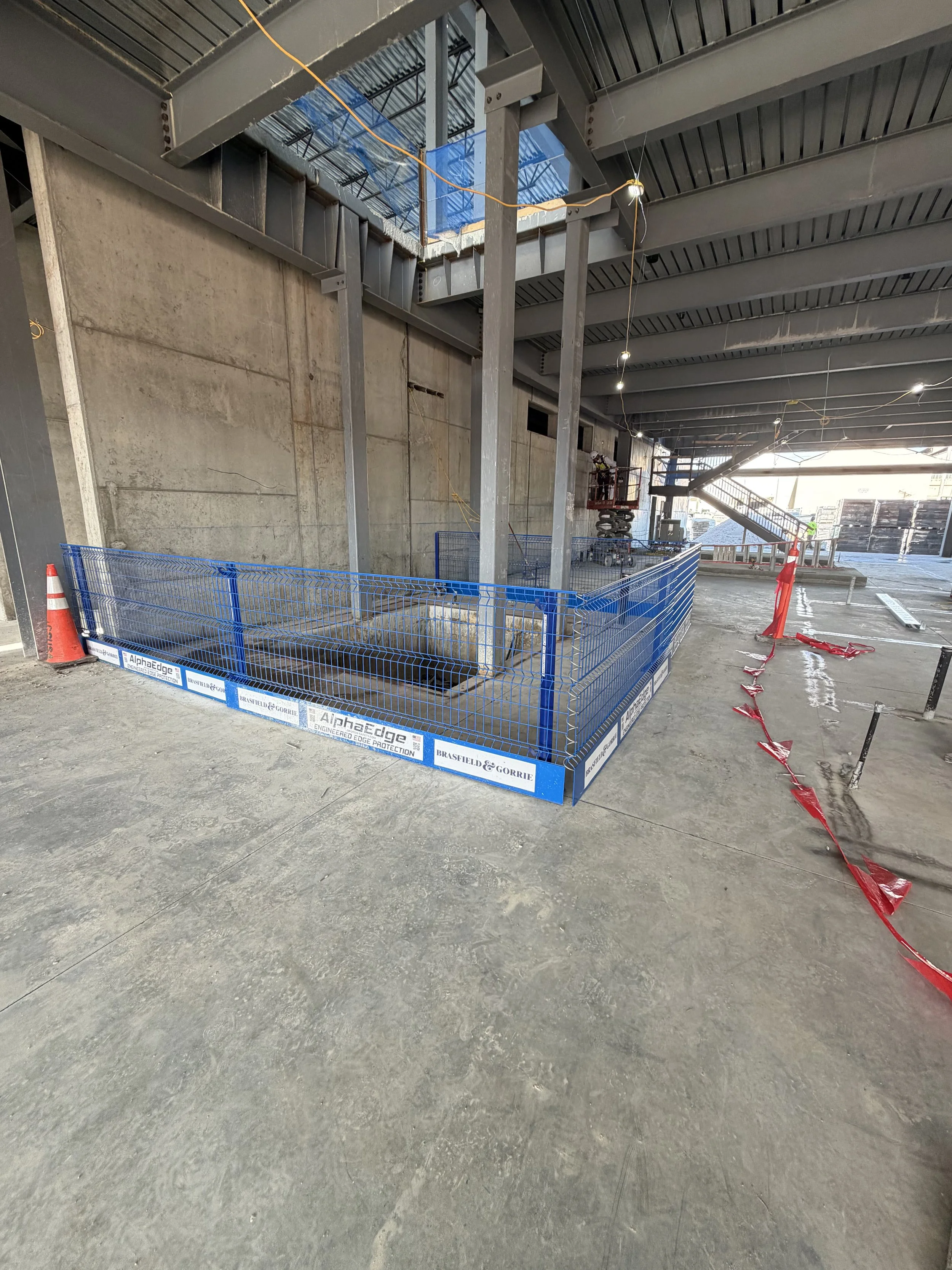 Construction site with a blue safety fence surrounding a large opening in the floor, with a worker on a lift nearby, and building framework overhead.