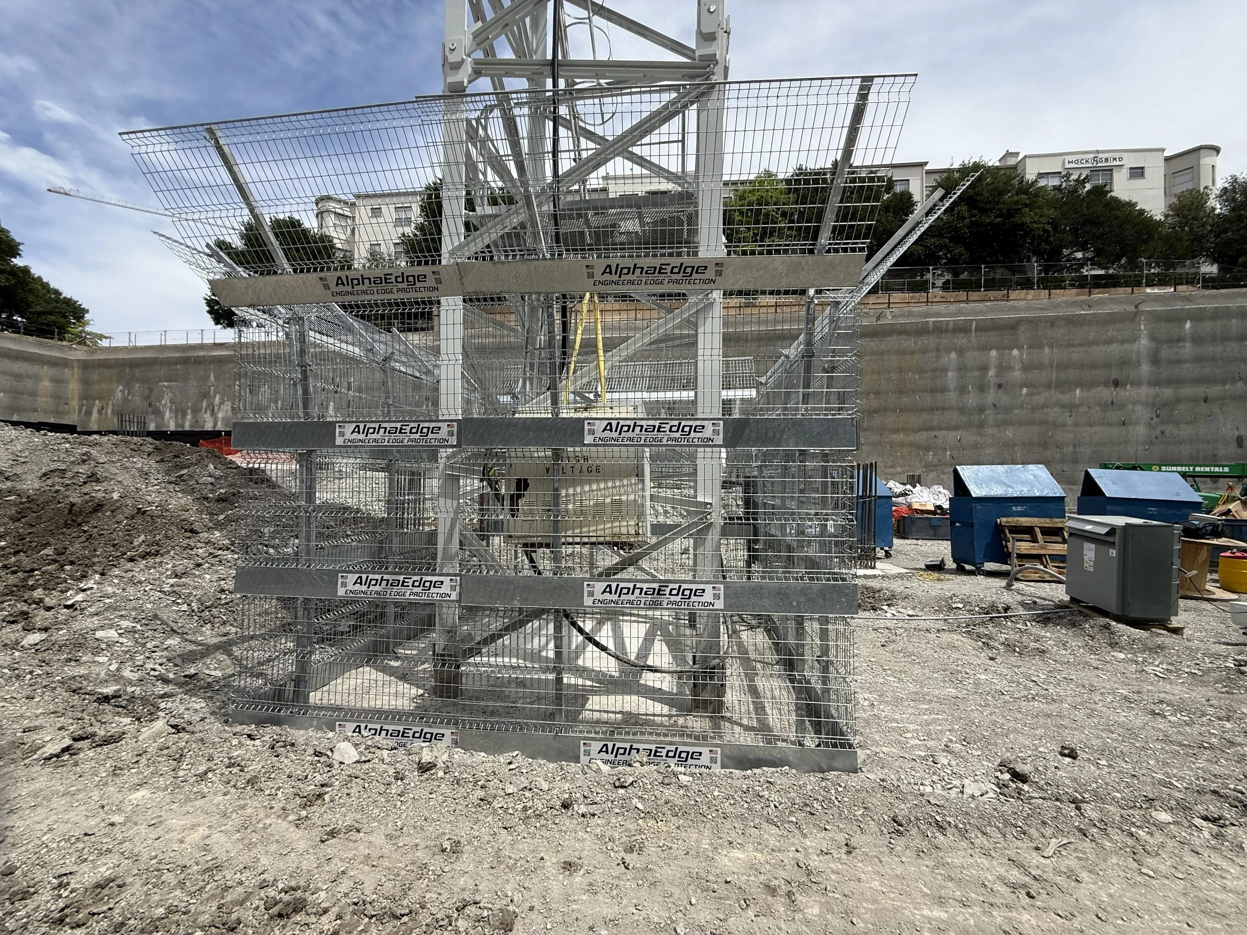Construction site with a large metallic tower or crane behind reinforced metal fencing with the brand name AlphaEdge. The ground is uneven dirt and gravel. In the background are trees and residential buildings under a partly cloudy sky.