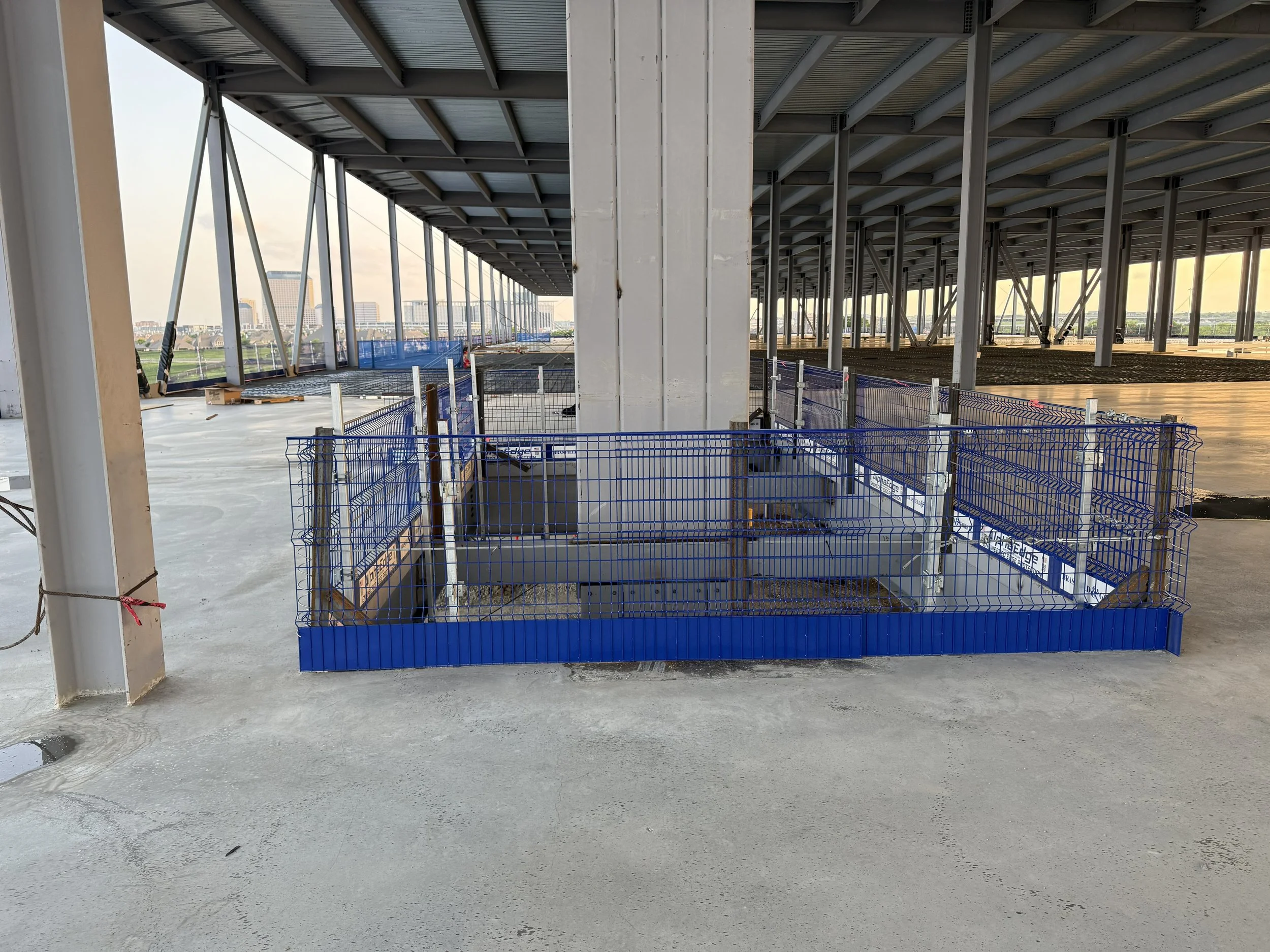 Construction site under a large roof structure with scaffolding and safety barriers, unfinished flooring, and a cityscape in the background.