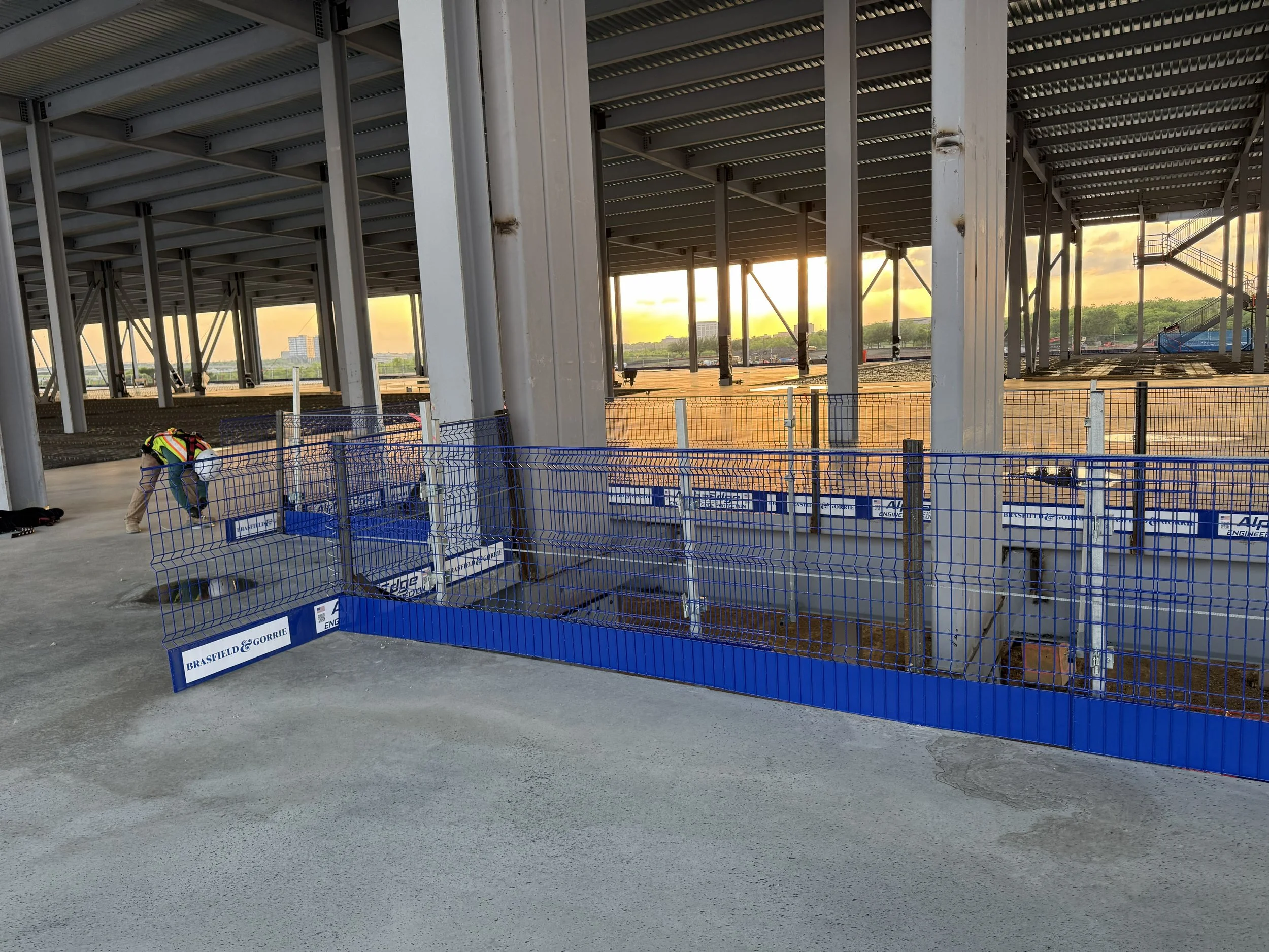 Construction site with steel framework, fencing, and person working, sunset sky in the background.
