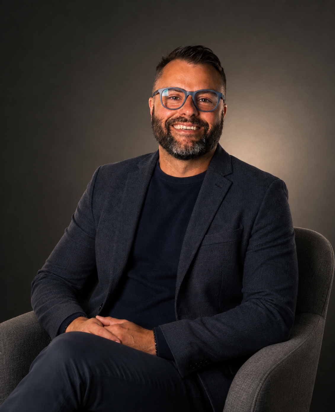 A man with glasses and a beard, wearing a dark blazer and black shirt, sitting in a gray chair against a gradient background, smiling at the camera.