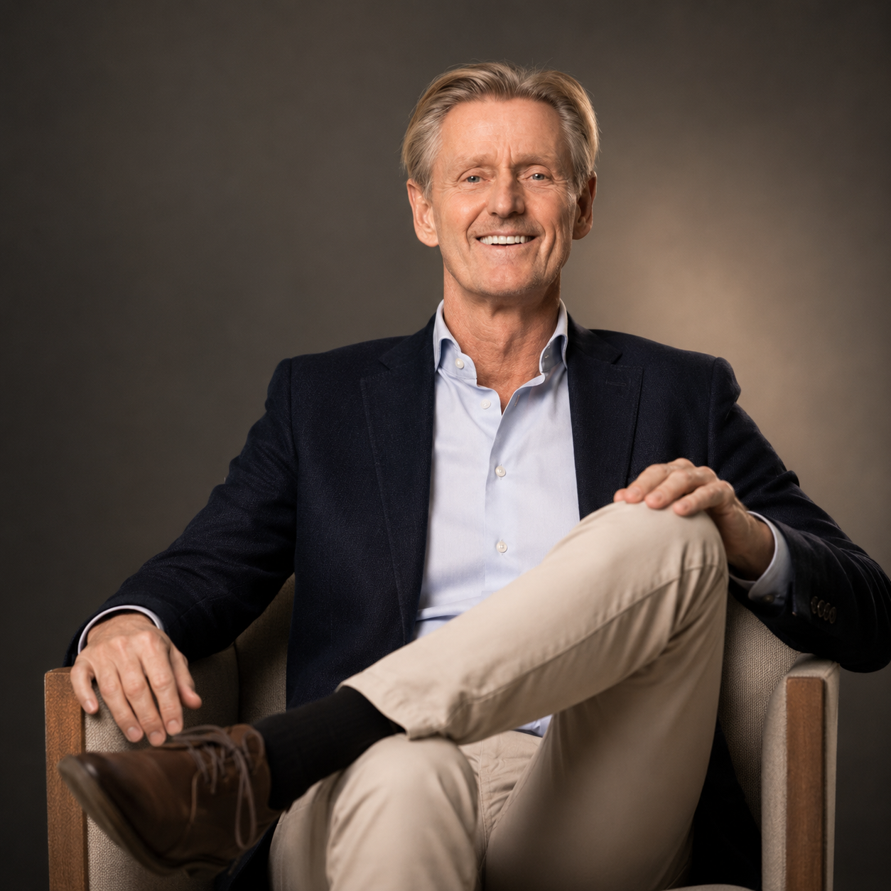 A smiling middle-aged man with gray hair, wearing a dark blazer over a light blue shirt, sitting comfortably in a beige armchair with his legs crossed, against a neutral background.