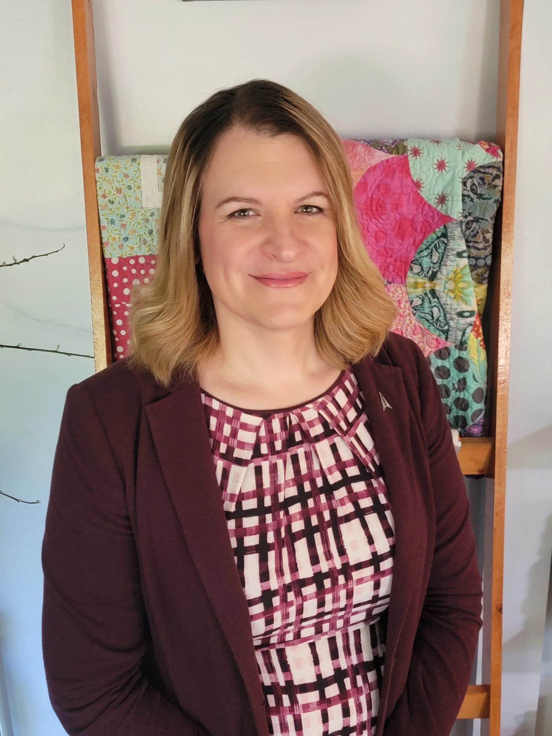 A woman with shoulder-length blonde hair wearing a maroon jacket and patterned top, smiling, standing in front of a colorful quilted wall hanging.