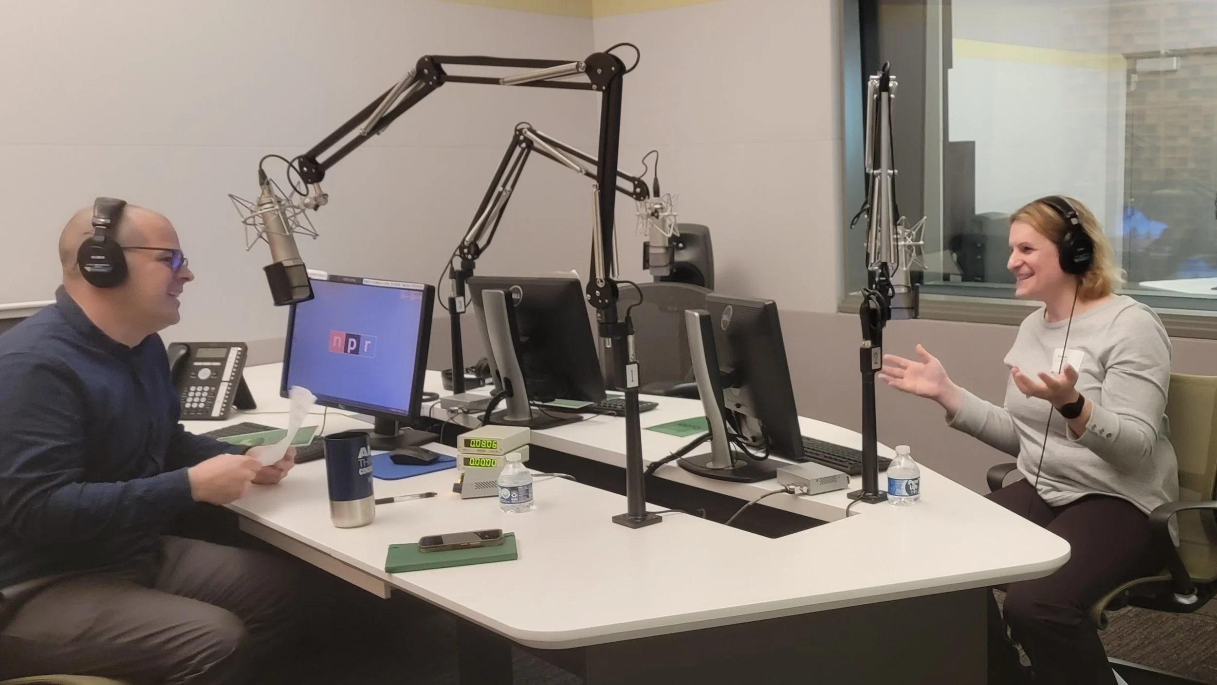 Two people with headsets are engaged in a conversation inside a radio broadcasting studio, with microphones and computer monitors in front of them.