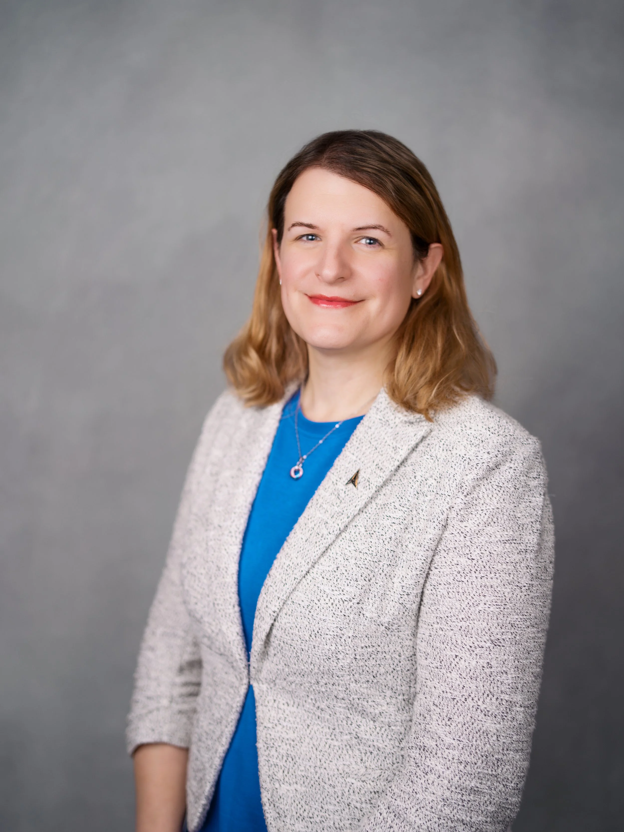 A woman with shoulder-length brown hair and blue eyes, smiling, wearing a blue top and a light-colored blazer, standing against a gray background.