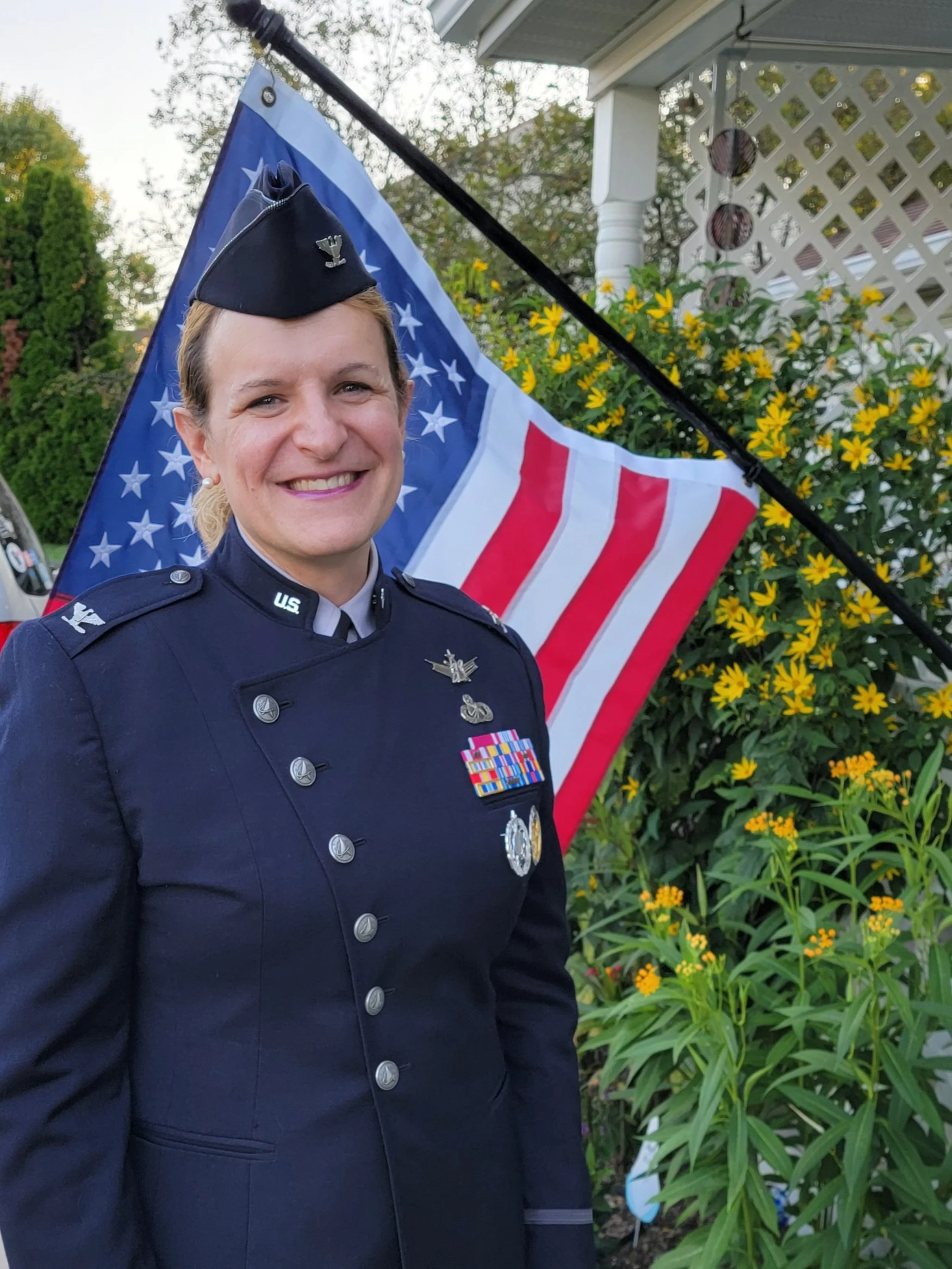 Close-up of a smiling woman in a United States Air Force uniform with medals and pins, standing in front of yellow flowers and American flag, outdoors.