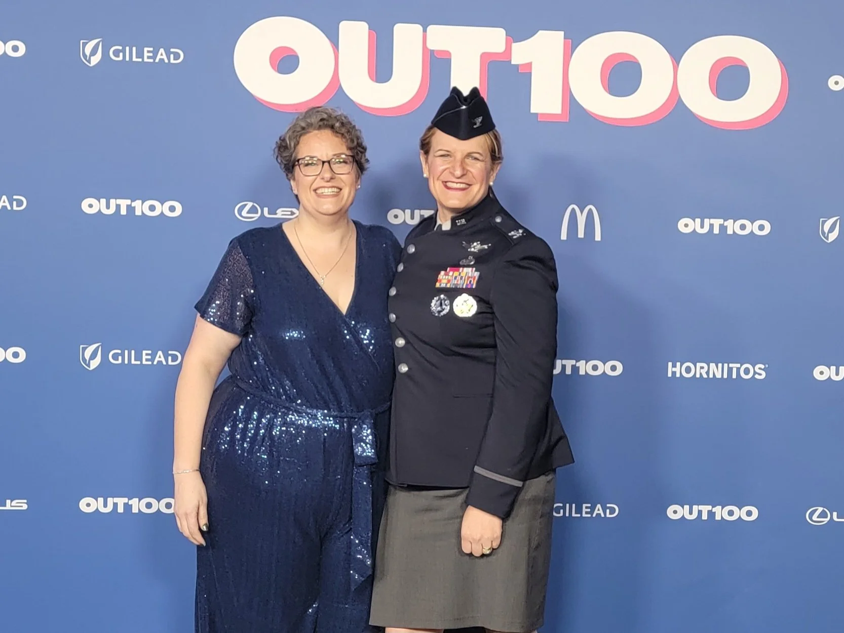Two women standing together at an event with a blue backdrop featuring logos and the text 'OUT100'. One woman is dressed in a sparkly dark blue dress with glasses, and the other in a military uniform with medals and badges, wearing a navy blue hat.