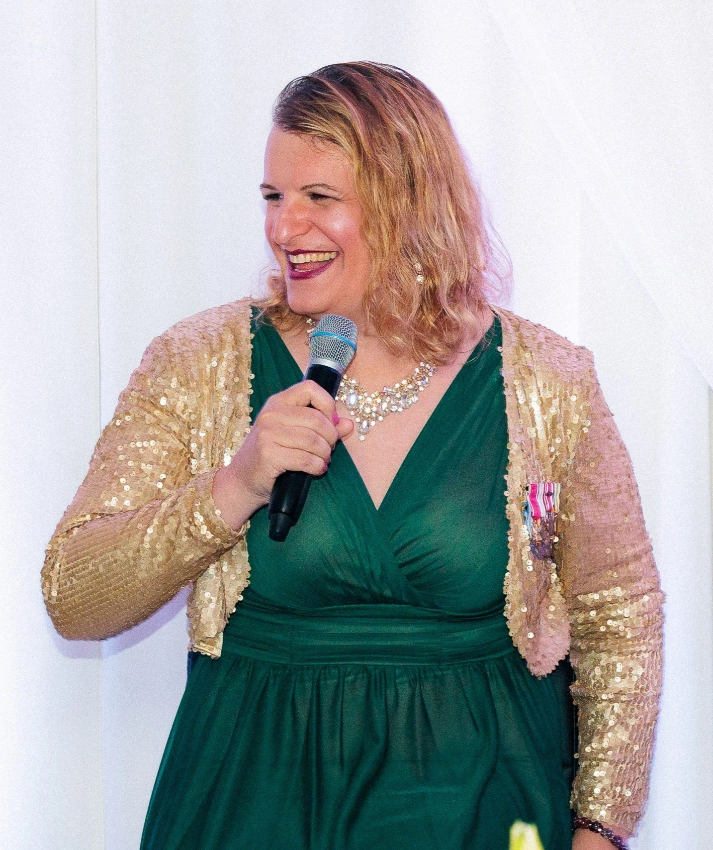 A woman with shoulder-length wavy hair, wearing a green dress, a gold sequin jacket, and a pearl necklace. She holds a microphone and is smiling.