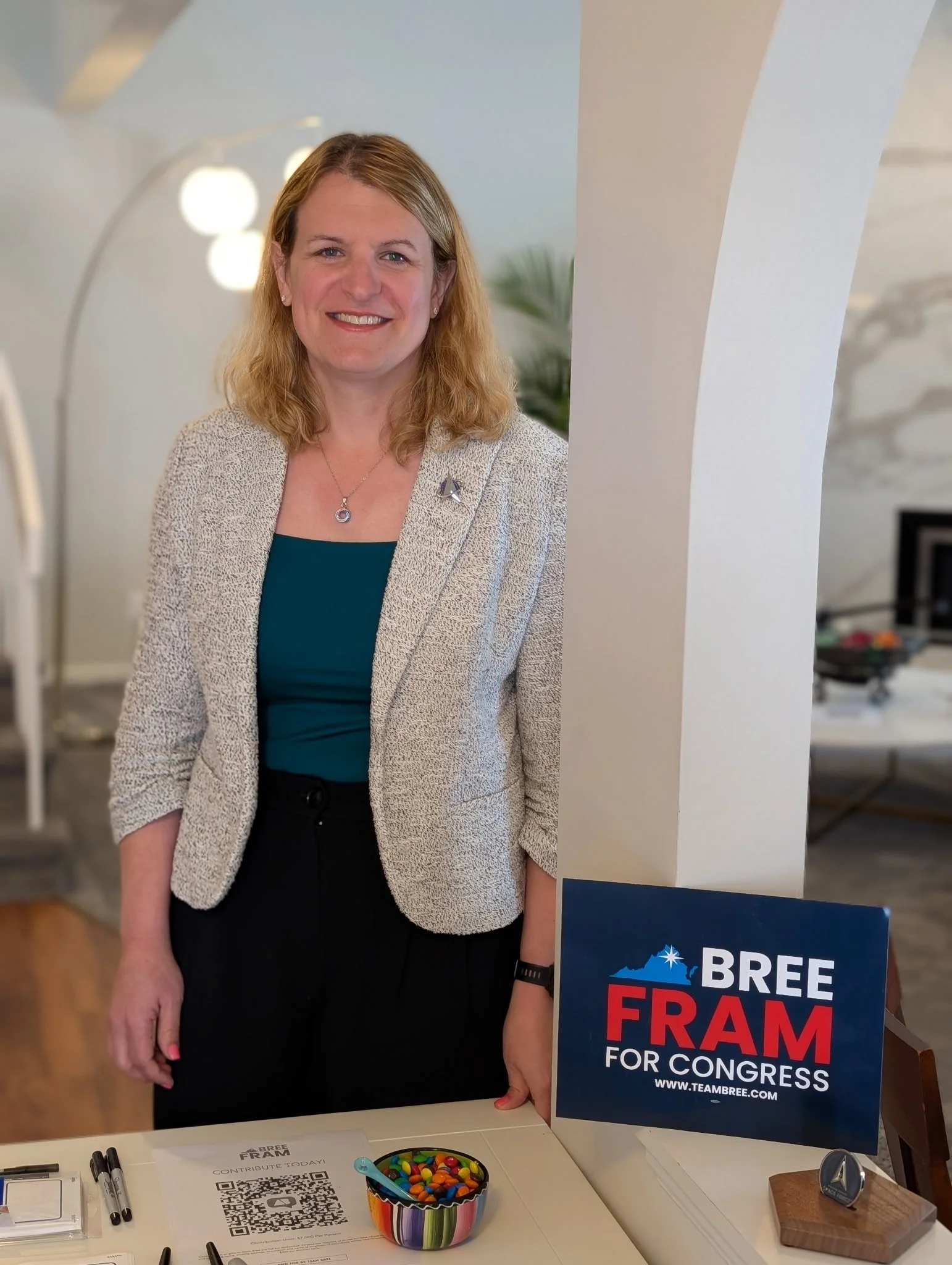 Bree Fram smiling at a desk with campaign signs reading "Bree Fram for Congress," a rainbow-colored candy dish, markers, and a QR code donation sheet.