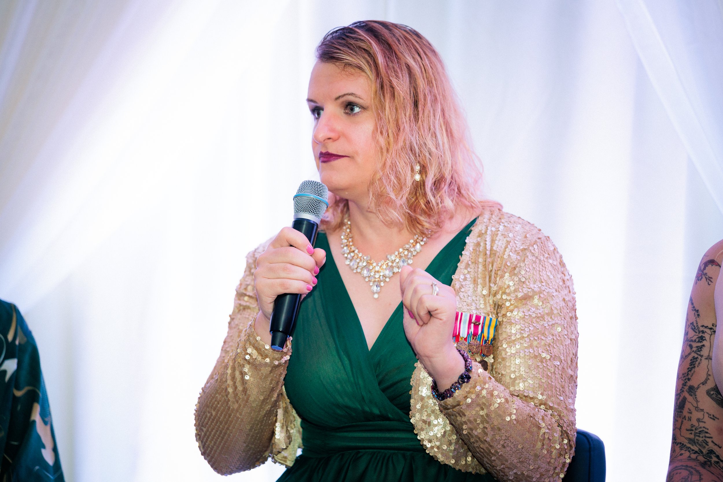 Woman with pinkish hair holding a microphone, wearing a green dress with a gold sequined jacket, and a necklace.