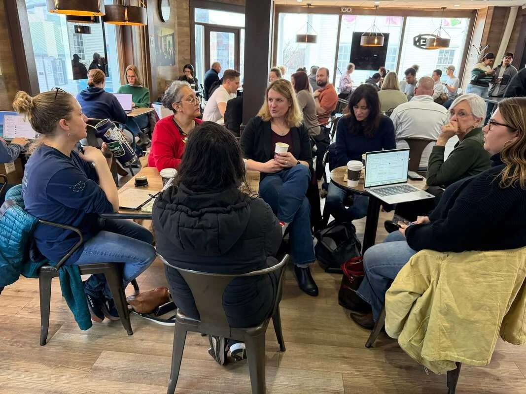 People sitting around tables in a busy coffee shop, engaged in conversation and working on laptops, with large windows and hanging lights overhead.