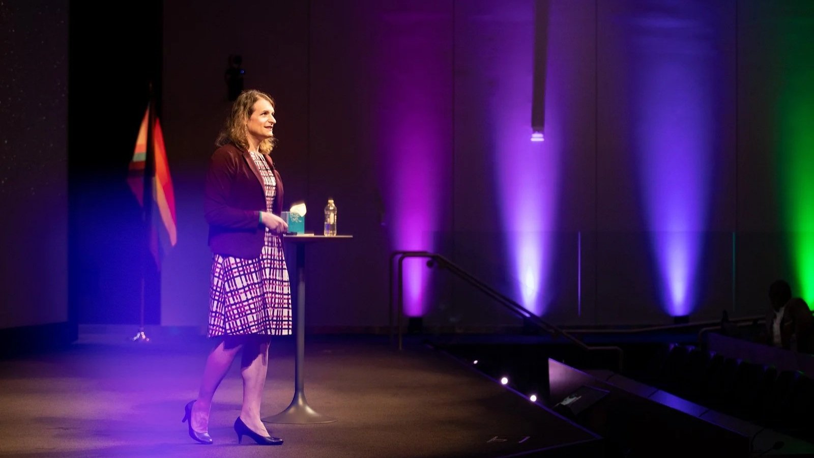 Woman standing on stage with purple and green lighting, holding a water bottle and a paper, next to a small table with a bottle of water and a box, in a dark auditorium.