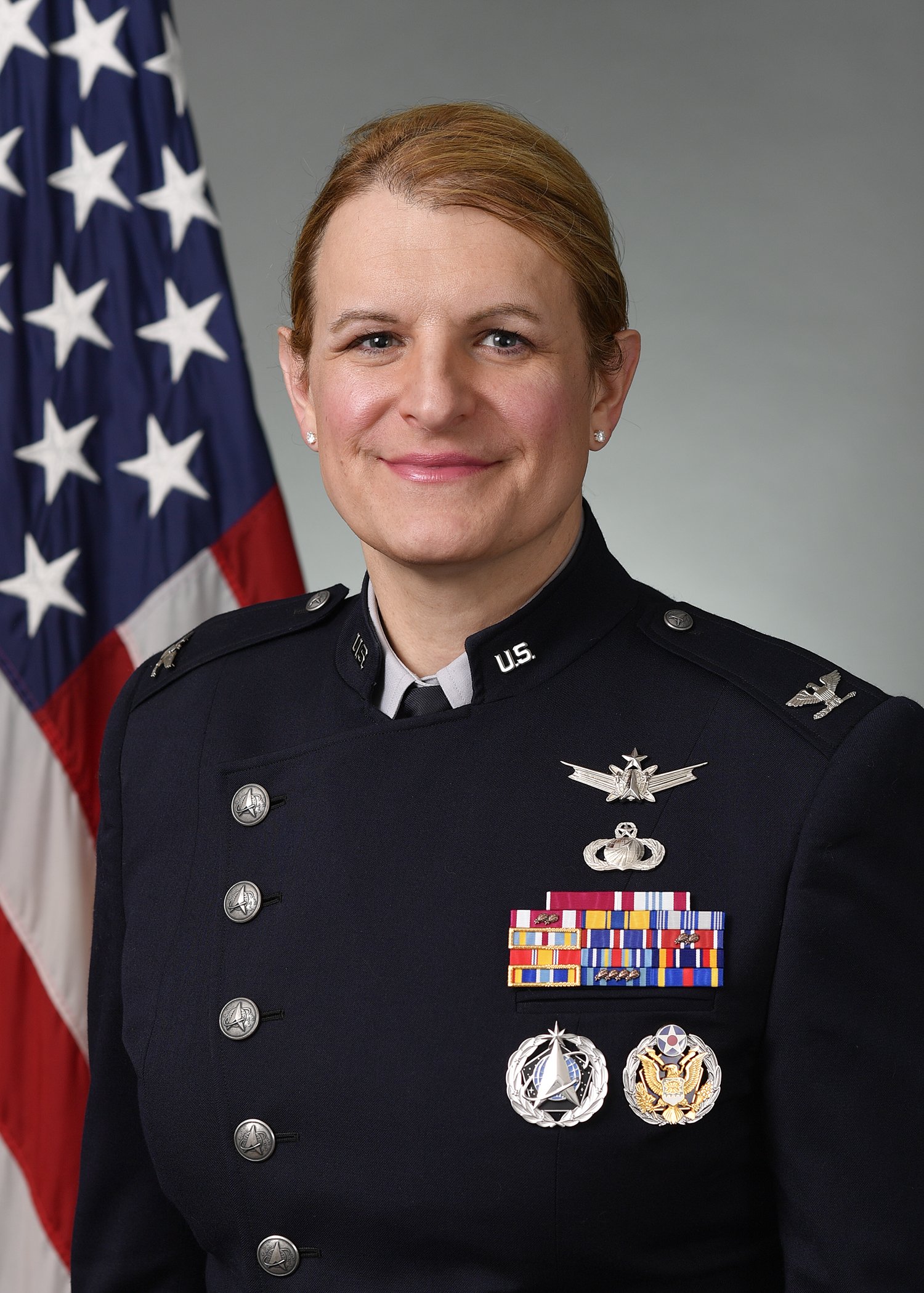 A woman in military uniform standing in front of an American flag. She is smiling, with ribbons and medals on her chest, and insignia on her shoulders and collar.