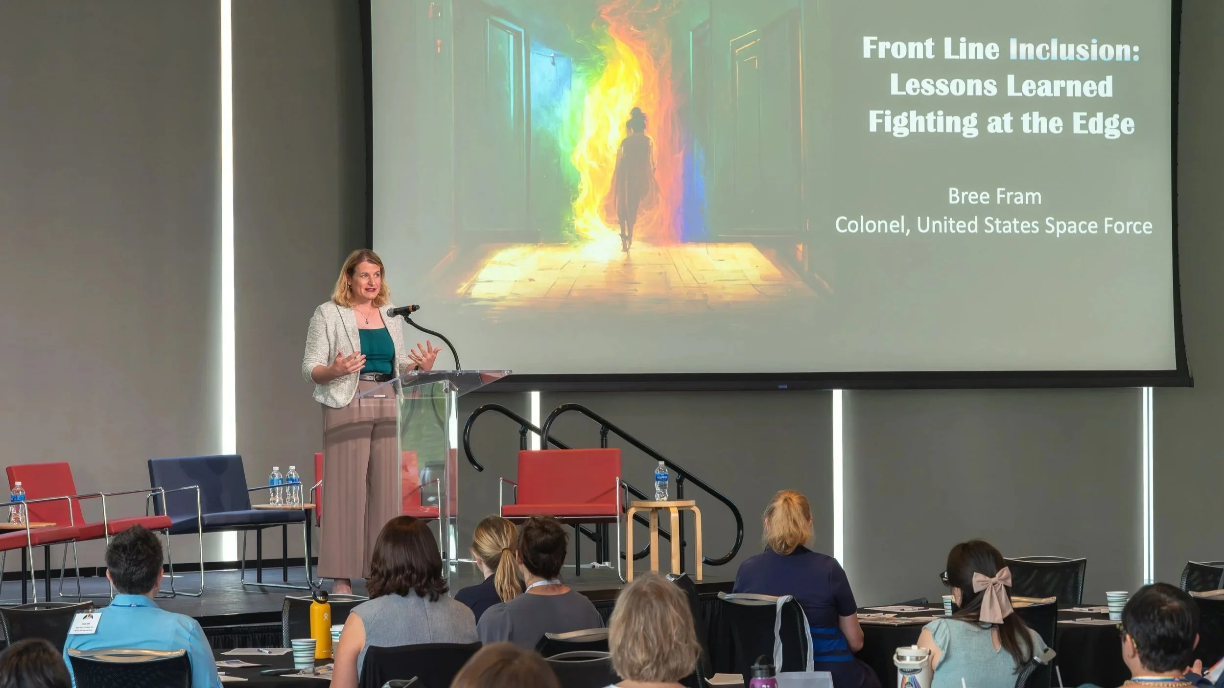 A woman presenting at a conference, standing at a clear podium, with a large screen behind her displaying a colorful abstract background and the presentation title "Front Line Inclusion: Lessons Learned Fighting at the Edge" and her name and title "Bree Fram, Colonel, United States Space Force." The audience, seated and facing her, is taking notes, and some have water bottles and cups on the tables.