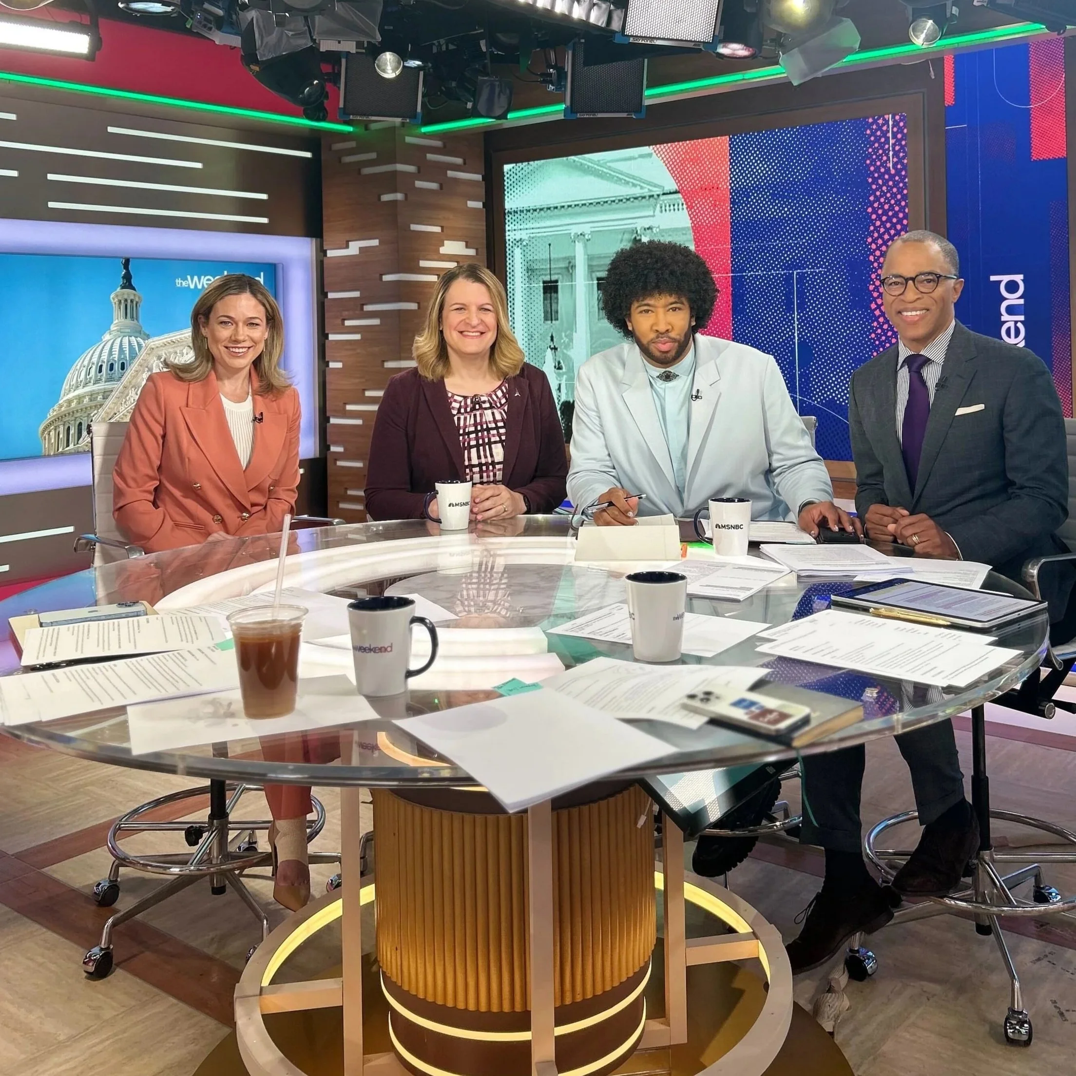 Group of four news anchors sitting at a news desk in a studio with a large digital screen and American government building background, papers scattered on the desk, coffee mugs, and various studio equipment overhead.