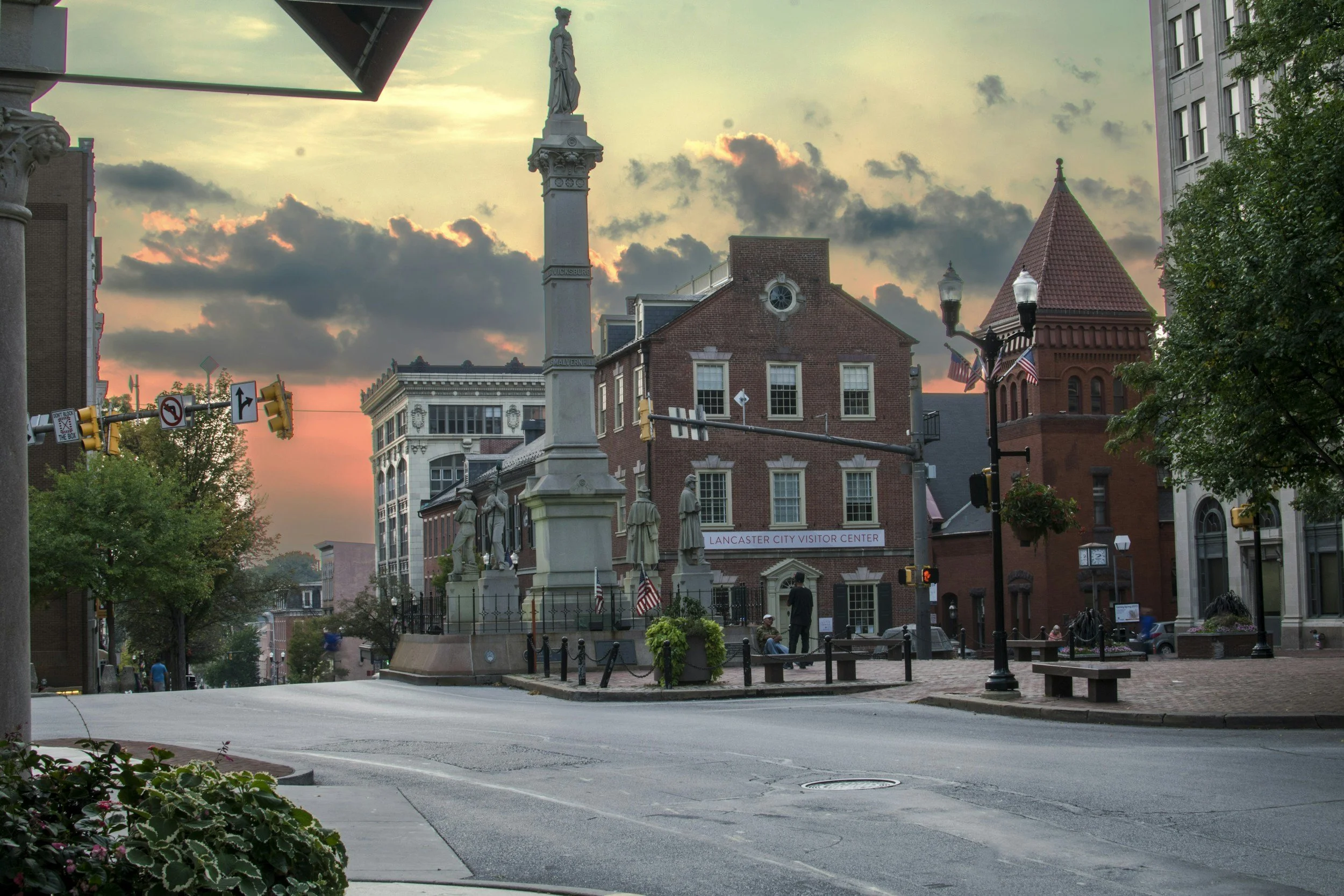 A city square at sunset with a tall monument topped by a statue of a woman, surrounded by historic brick buildings, trees, street lamps, American flags, and people walking.