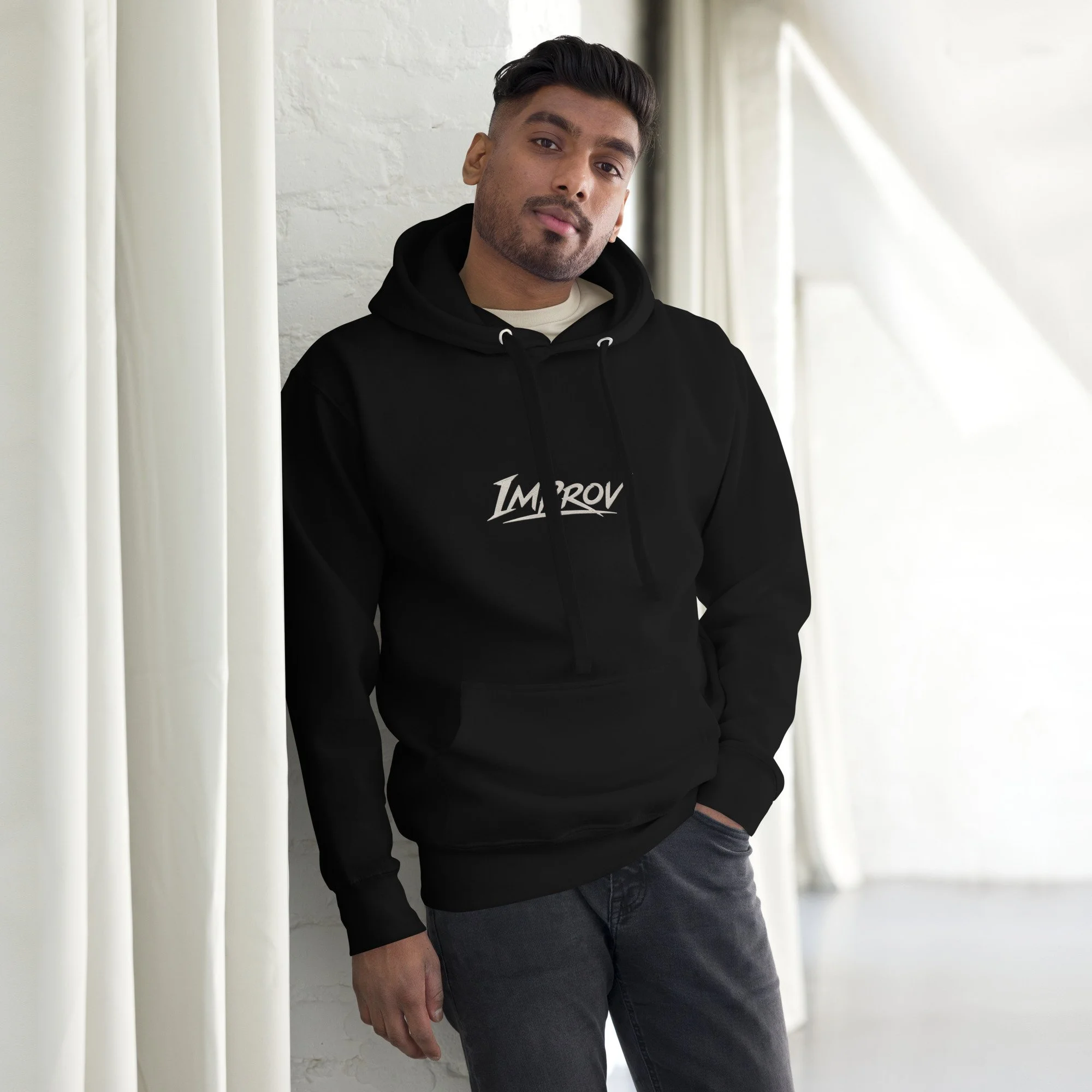 A young man with dark hair and beard leaning against a white wall with vertical blinds nearby, wearing a black hoodie and jeans.