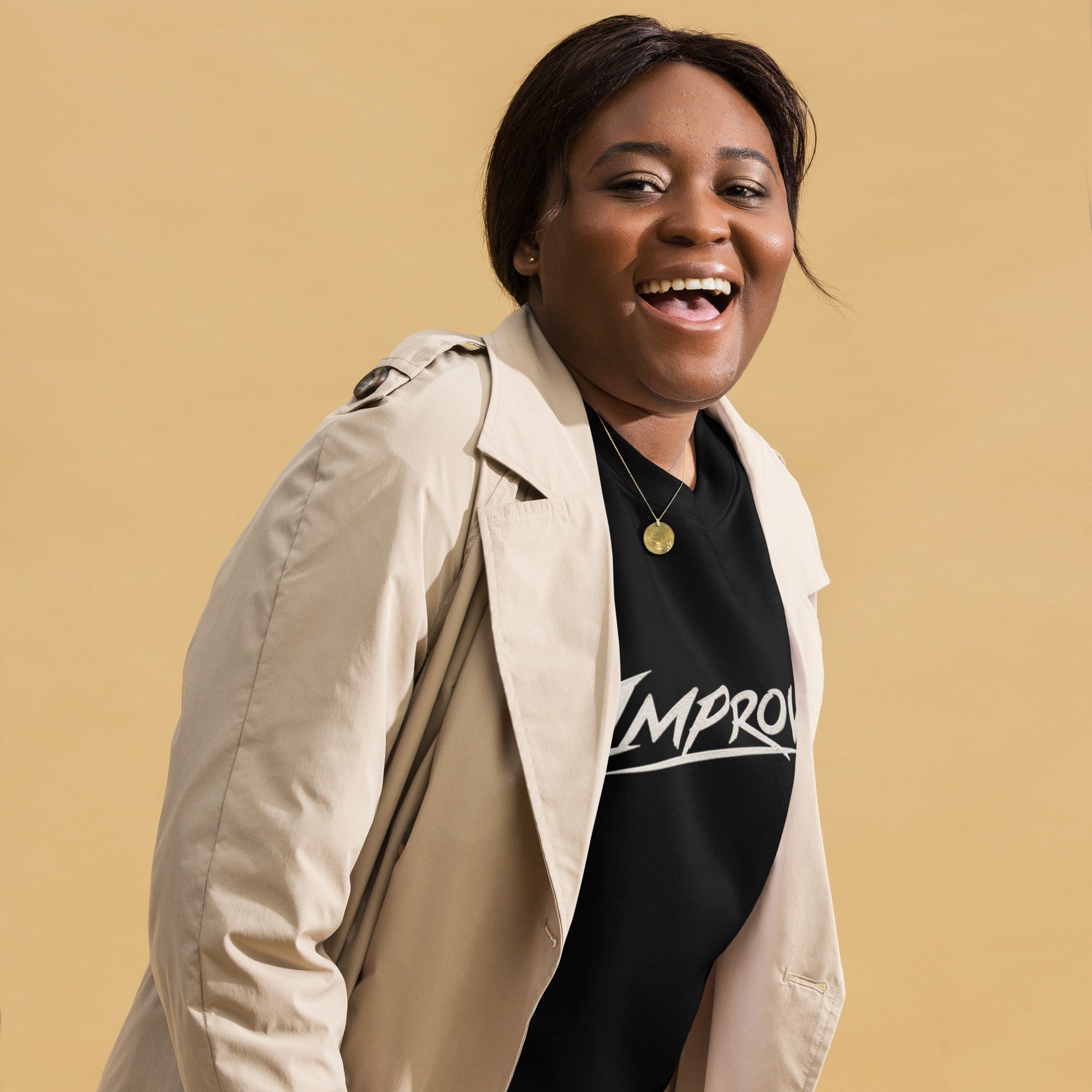 Close-up of a woman with short dark hair, smiling and showing her teeth, wearing a black T-shirt with white text, a beige trench coat, and a gold pendant necklace, against a plain beige background.
