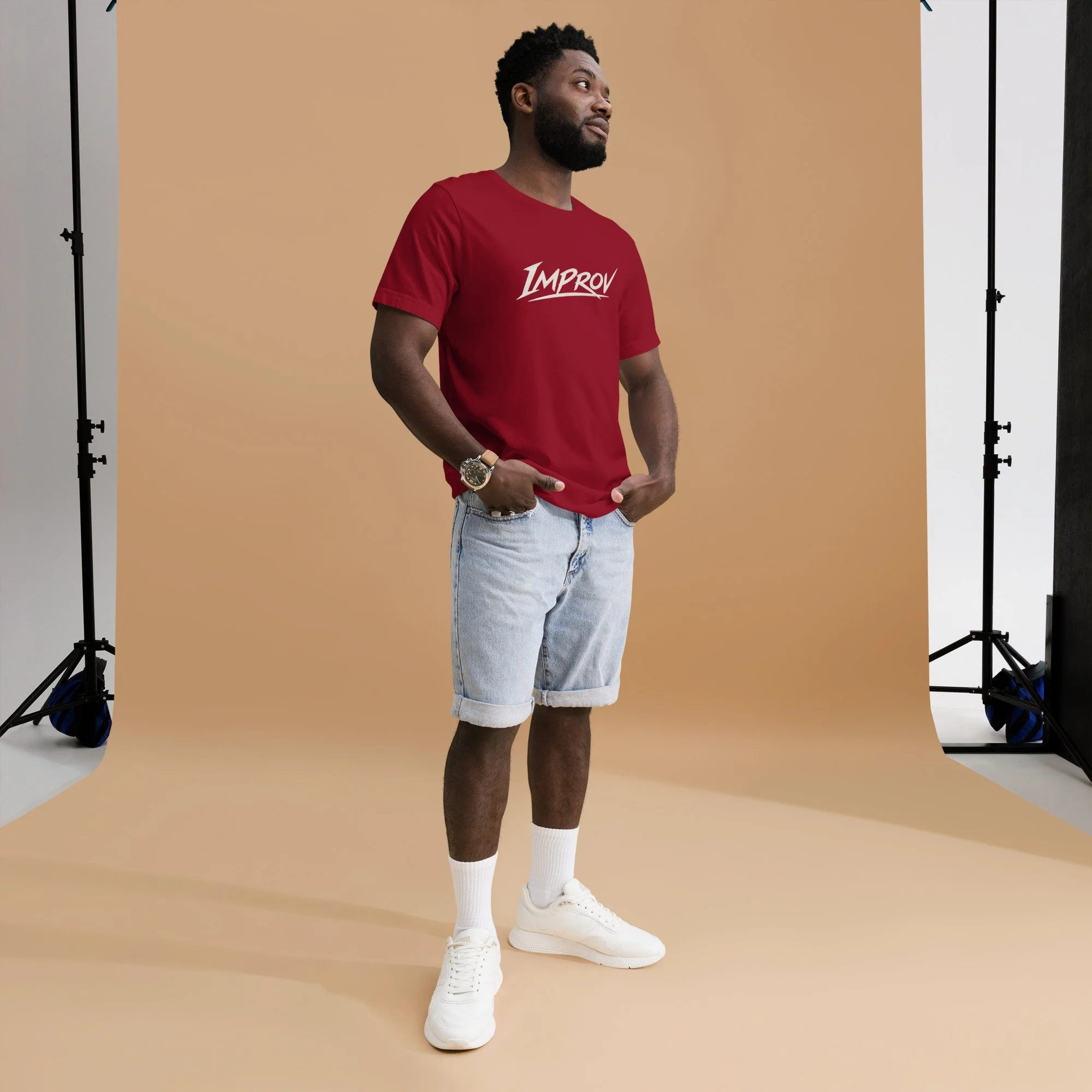 Young man with a beard wearing a red T-shirt with 'IMPROV' written on it, light blue jeans, white sneakers, and white socks, standing in front of a beige background during a photo shoot.