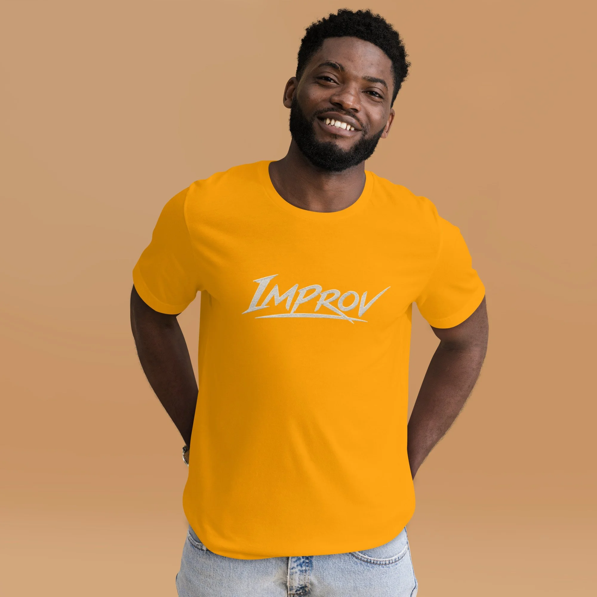 Smiling African American man with a beard and short curly hair, wearing a yellow T-shirt with the words "I'M PROV" printed on it and blue jeans, standing against a beige background.