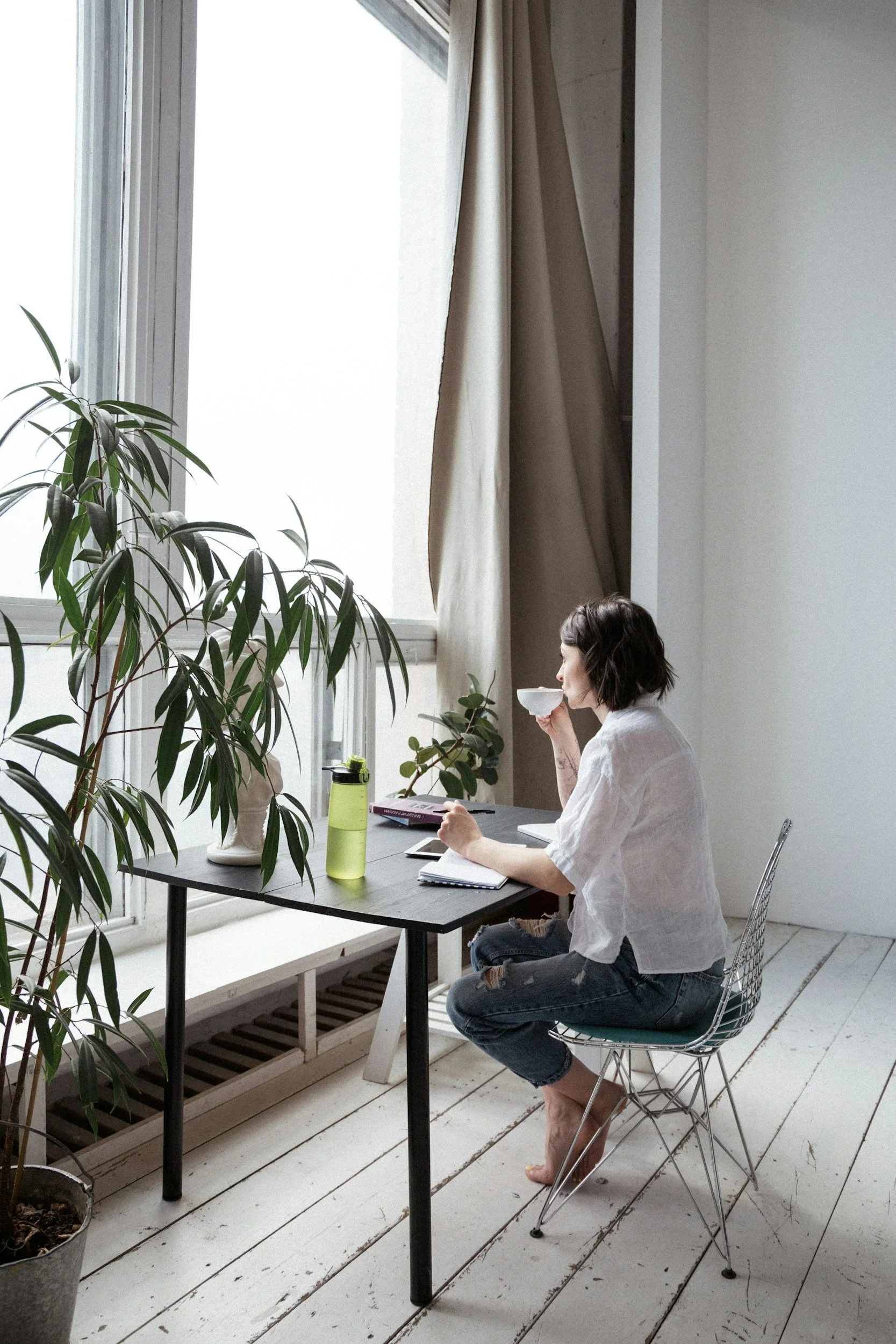 A woman sitting at a table by a large window, drinking from a white mug. The room has white walls, wooden flooring, and various plants on and near the table. The woman is wearing a white shirt and ripped jeans, and there are notebooks, a tablet, and a green water bottle on the table.