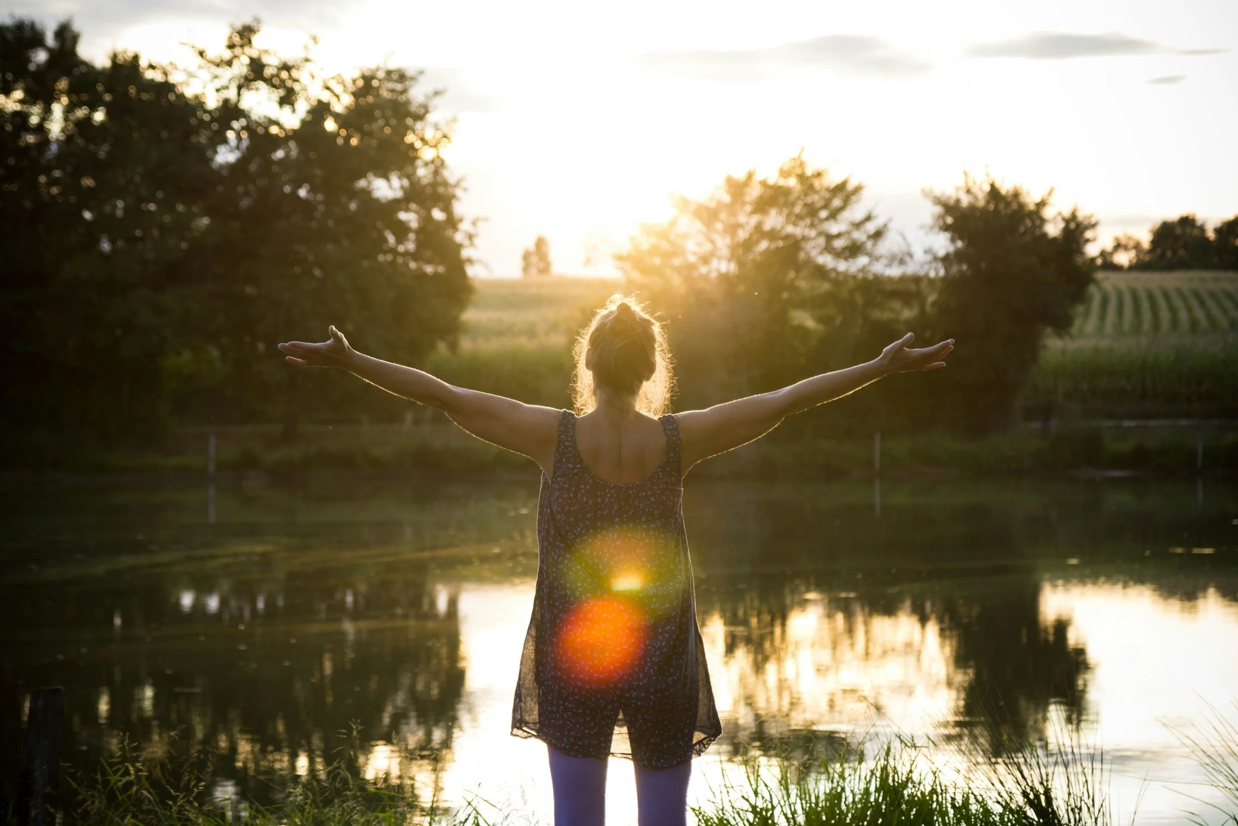 A woman standing by a lake with her arms outstretched during sunset, surrounded by trees and a grassy area.