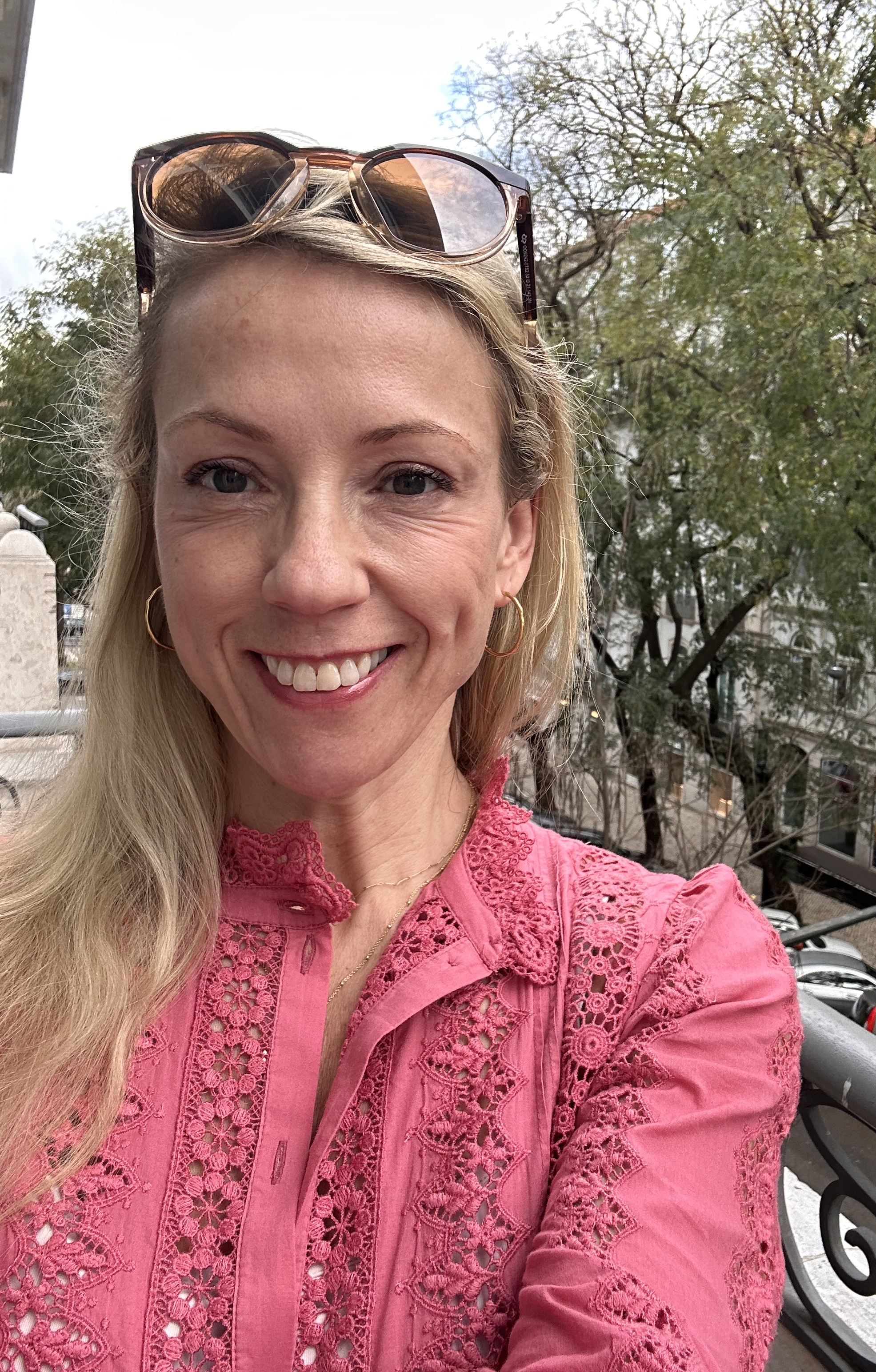 A smiling woman with blonde hair and sunglasses on her head is wearing a pink embroidered blouse and taking a selfie outdoors on a balcony with trees in the background.