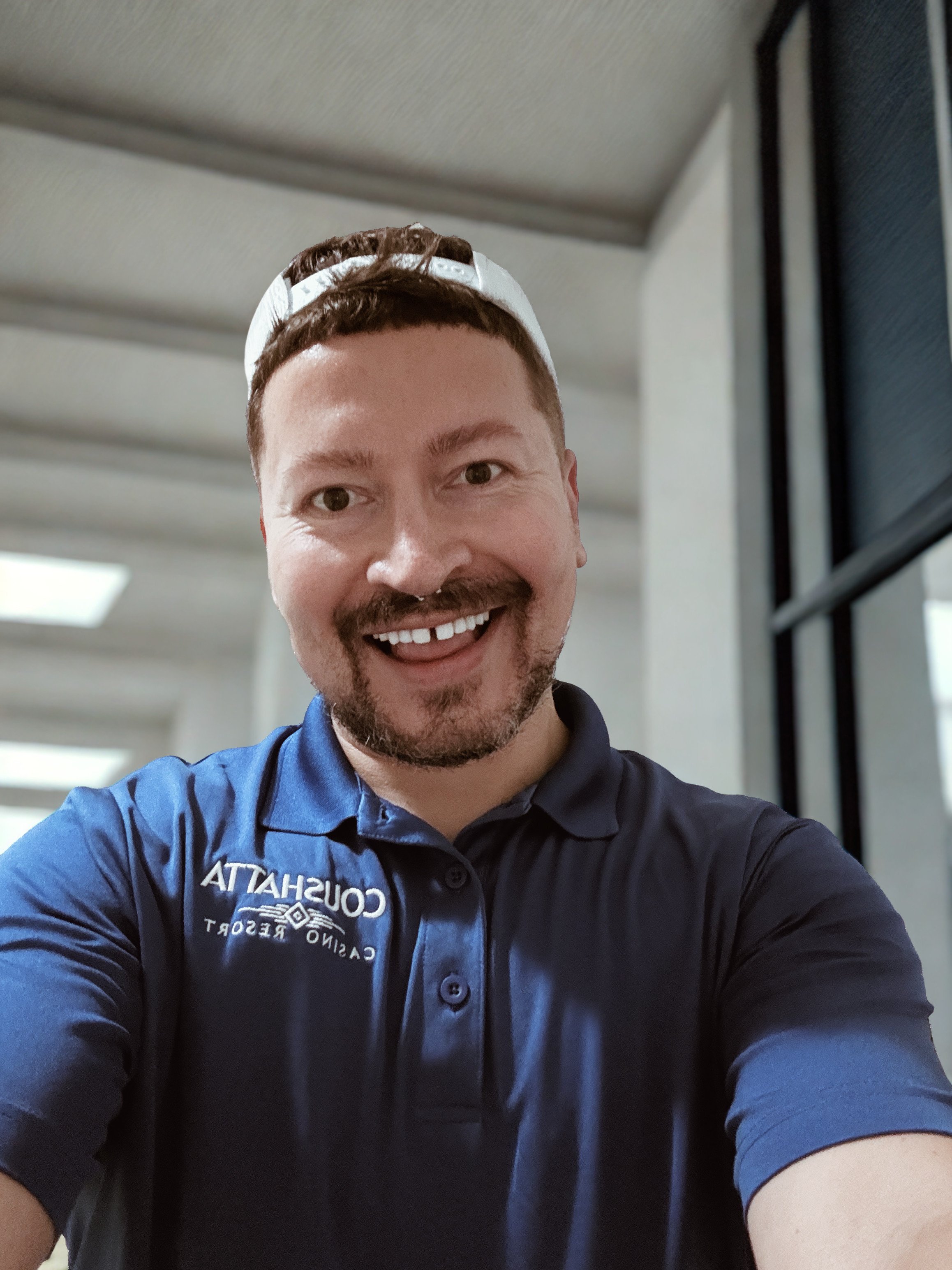 A man with light skin, short brown hair, a goatee, and a mustache, smiling at the camera in a selfie. He is wearing a blue shirt with a logo and text, and a white cap worn backward. The background is blurred with indoor ceiling lights and windows.