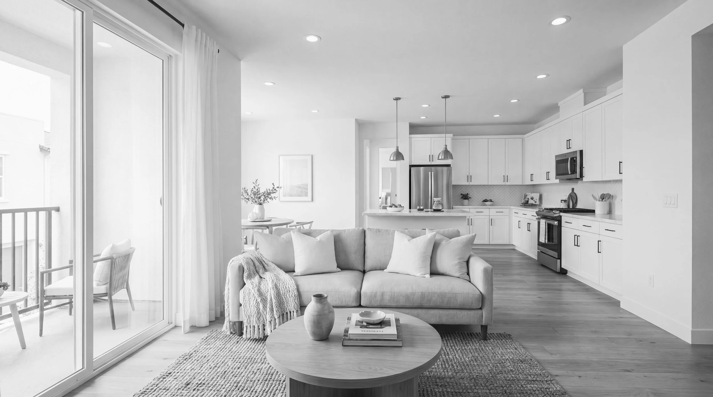 Black and white photo of an open-concept living room and kitchen area with modern decor, a sliding glass door leading to a balcony, a sofa with pillows and a throw blanket, a round coffee table with vases and books, and white kitchen cabinets with stainless steel appliances.