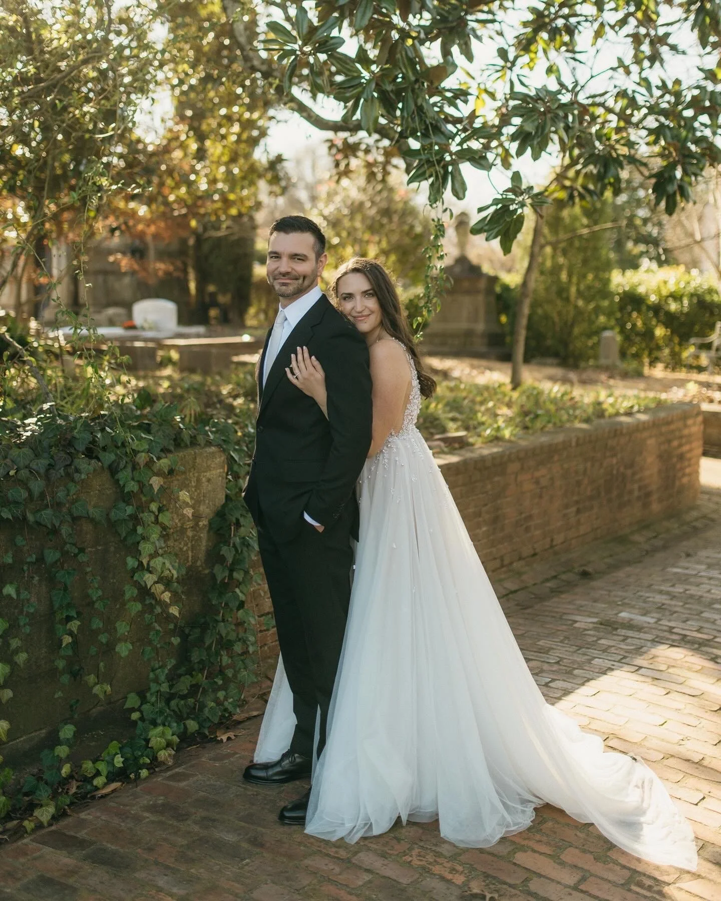 A bride and groom standing close together outdoors on a brick pathway, surrounded by trees and foliage, with the bride embracing the groom from behind. The groom is dressed in a black tuxedo with a white shirt and tie, smiling slightly. The bride is in a white wedding gown with lace details, smiling gently. The sunlight filters through the trees, creating a warm, golden atmosphere.