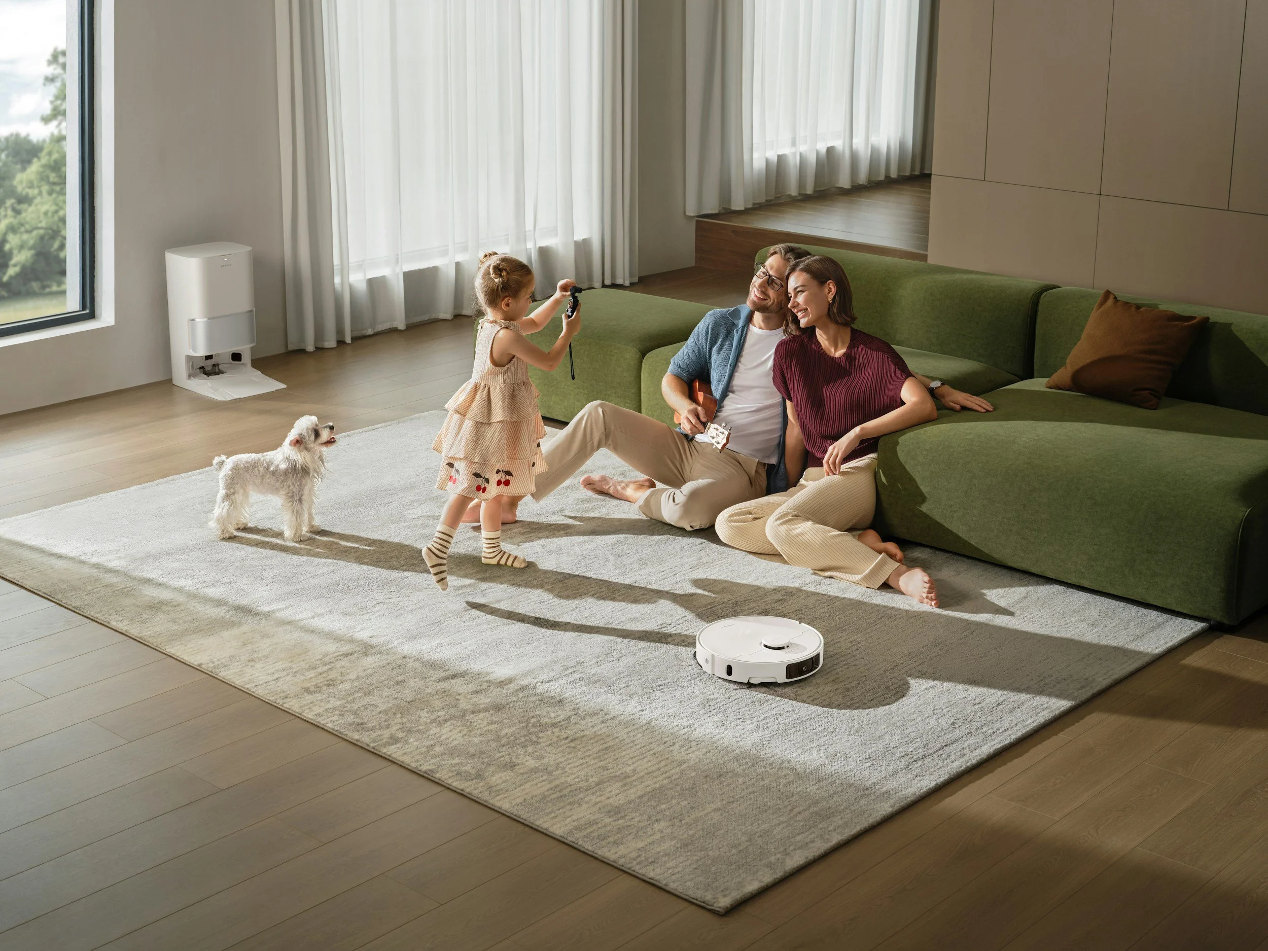 A family enjoying time together at home: a man and woman sitting on a green sofa laughing, a young girl standing holding a toy, and a small dog, with a robotic vacuum cleaner on the floor.
