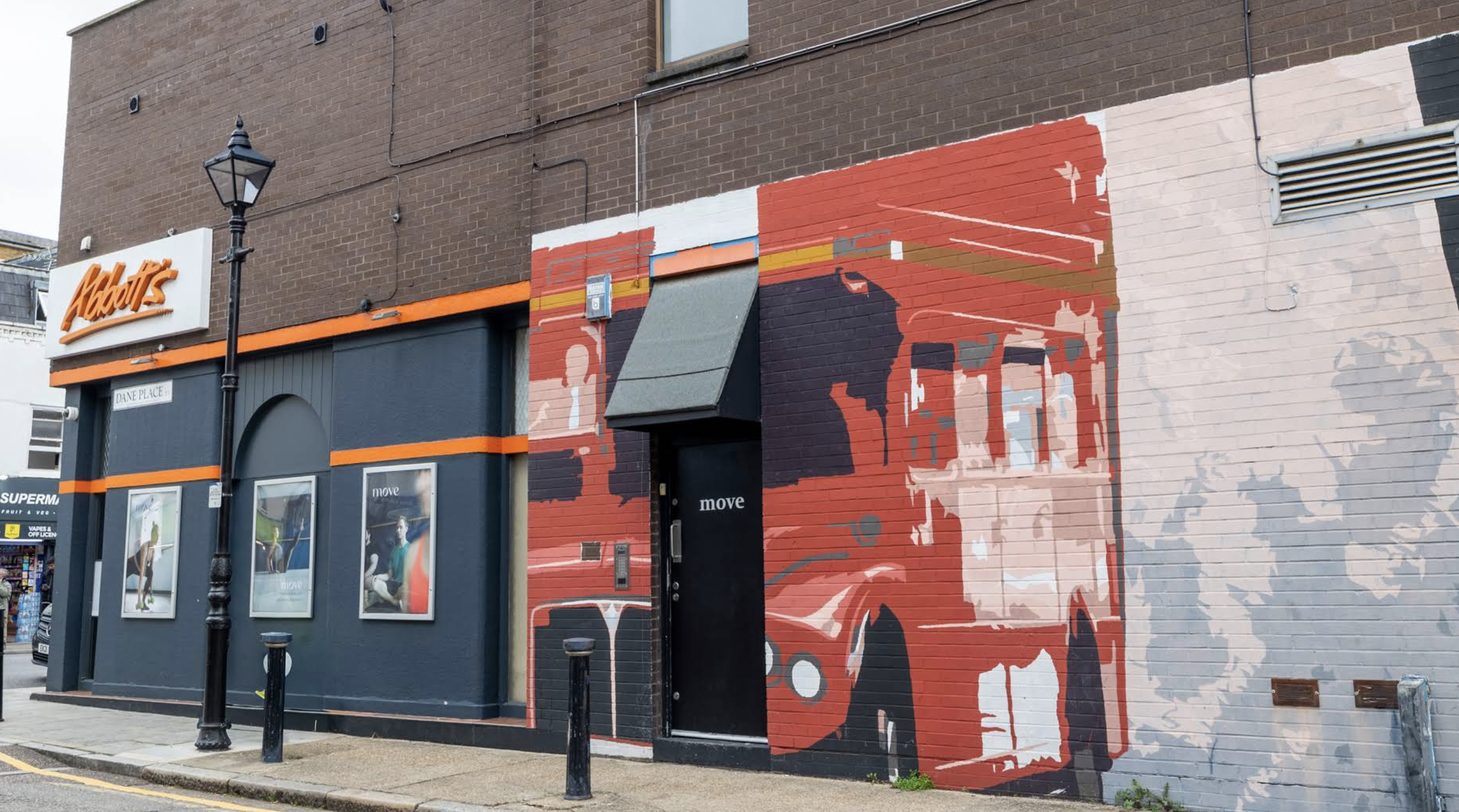 Street entrance of Move Studio London on Dane Place, featuring a large mural, a black street lamp, window posters and an Abbot’s sign next to the entrance.