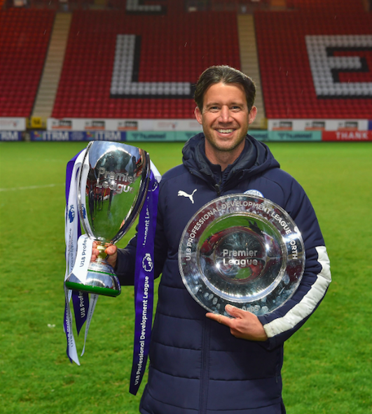 A man smiling and holding two trophies on a football field, with empty stadium seats in the background.