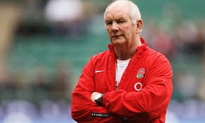 An elderly man wearing a red sports jacket with a logo, standing with arms crossed, outdoors at a sports venue.