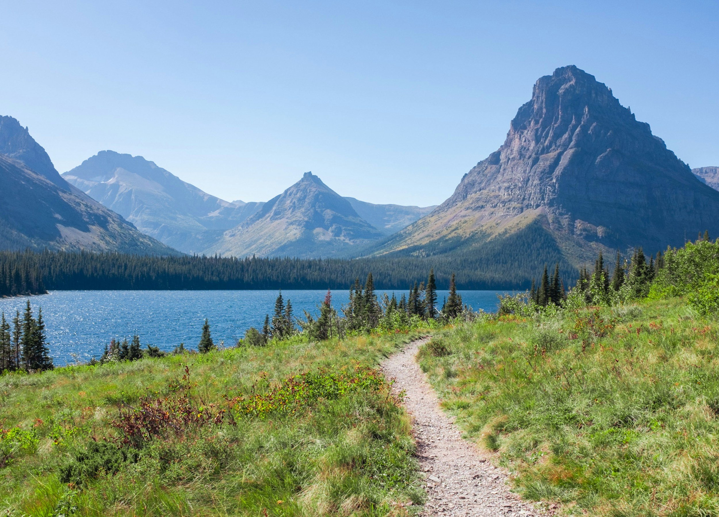 A scenic view of a mountain landscape with a trail leading through green grass, a lake, and rugged mountains in the background under a blue sky.