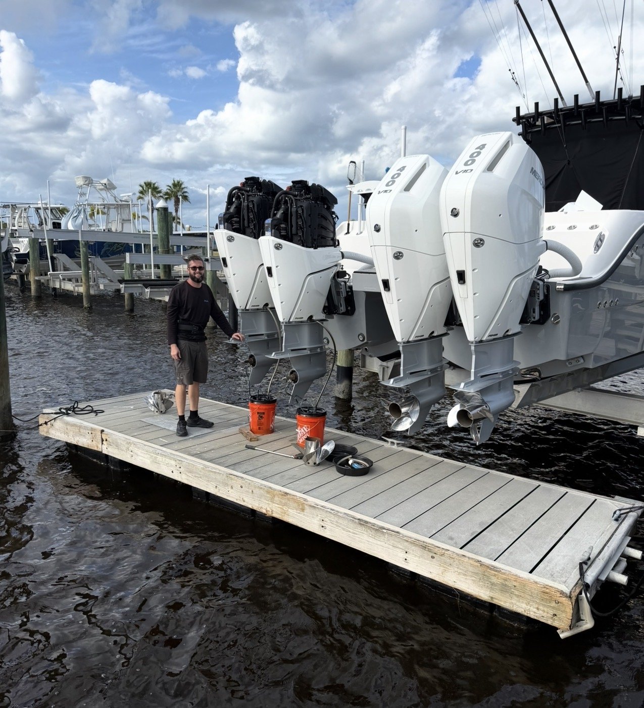 Man standing on a wooden dock with two orange buckets, a wrench, and other tools, next to a large boat with three outboard motors, at a marina with boats and palm trees in the background.