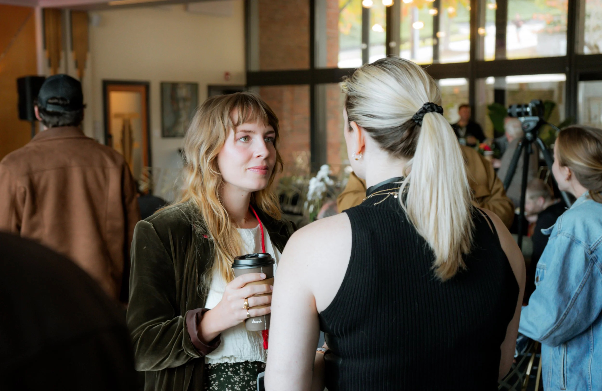 Two women are engaging in conversation at a social event inside a building with large windows. One woman with blonde hair tied back is holding a coffee cup, while the other woman has wavy light brown hair. The background shows other people, a camera, and indications of a casual or professional gathering.