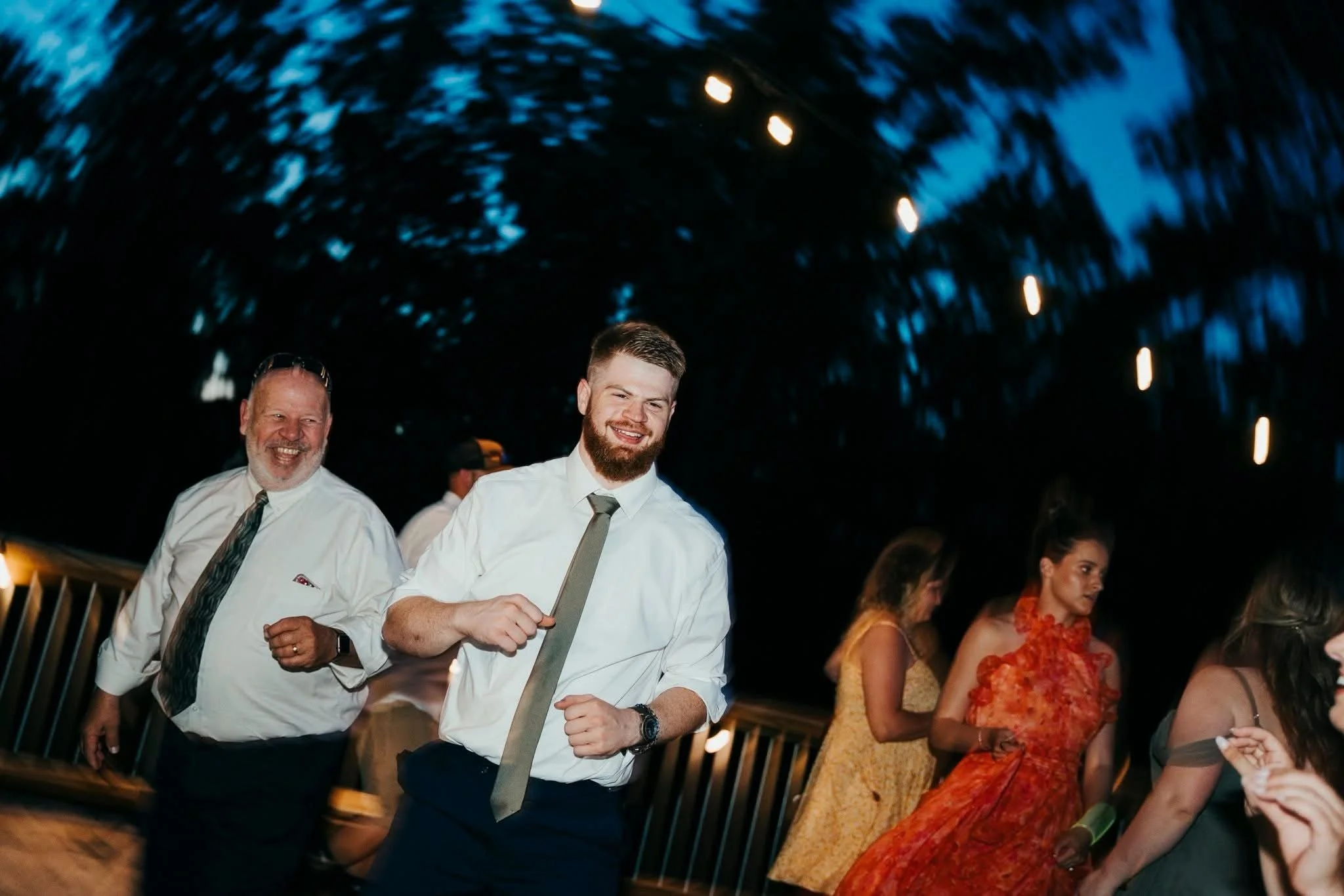 Smiling young man in white shirt and tie dancing at a party or celebration, surrounded by other people, with blue string lights hanging above.