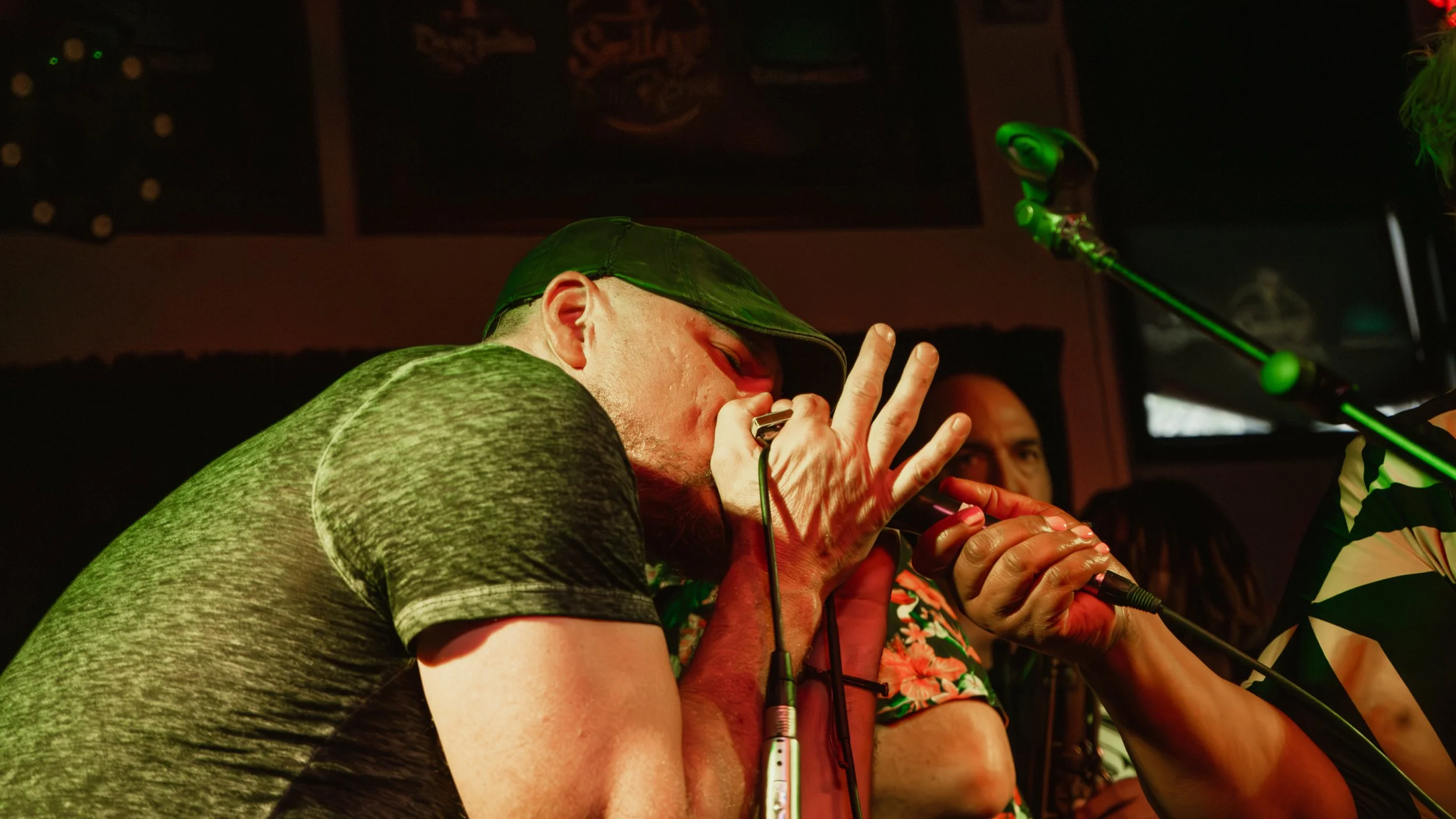 People singing into microphones in a dimly lit setting, with one person wearing a gray cap and gray clothing, and green lighting effects.