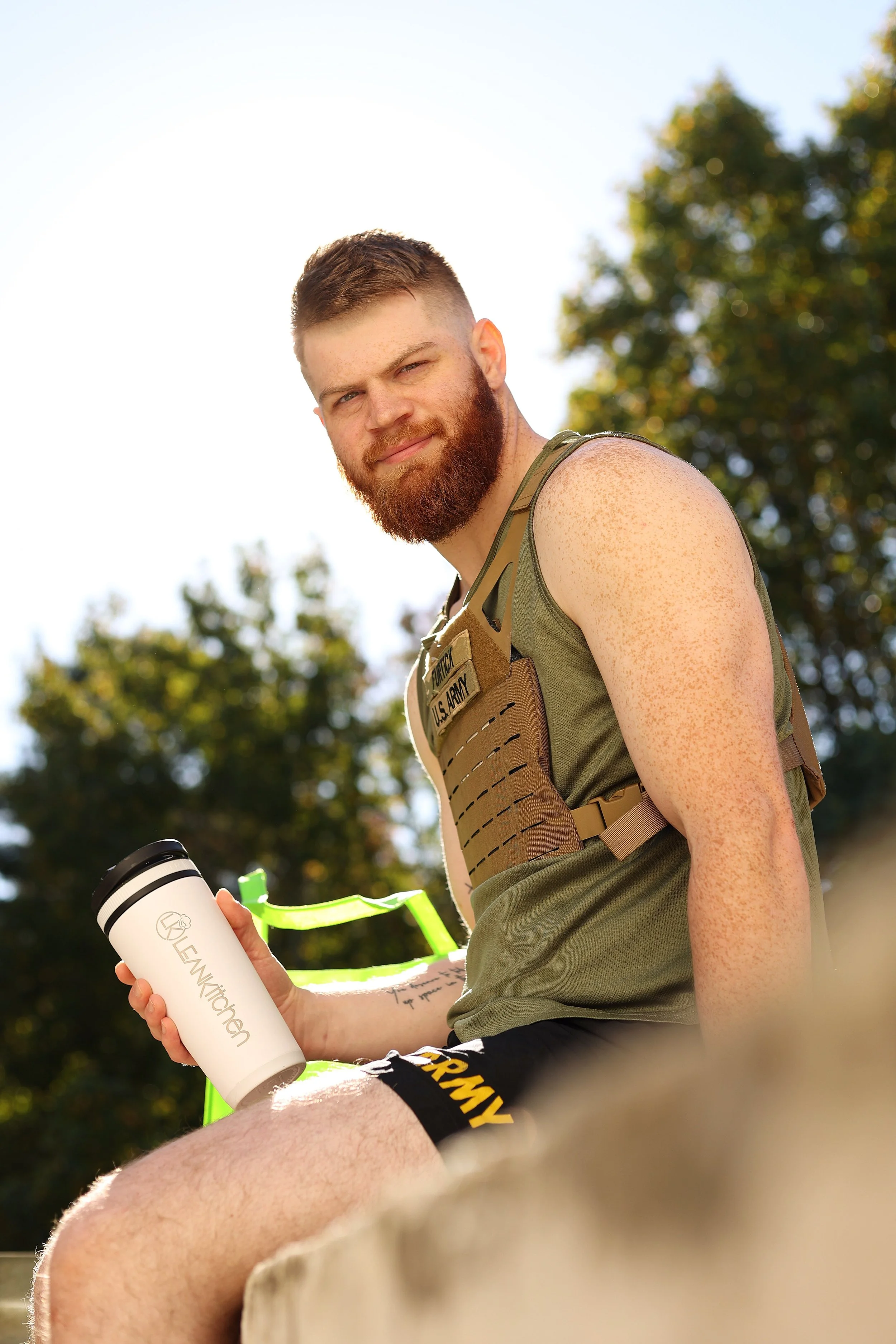A young man with a beard and freckles, wearing a green tank top and black shorts with yellow lettering, sitting outdoors on a sunny day. He is holding a white water bottle with the words Lean Kitchen on it.