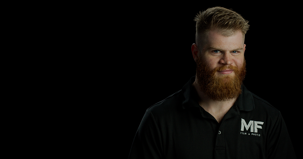 Portrait of a man with a red beard and short, styled hair wearing a black polo shirt with a logo that reads 'MF Film & Photo' on the chest, against a black background.